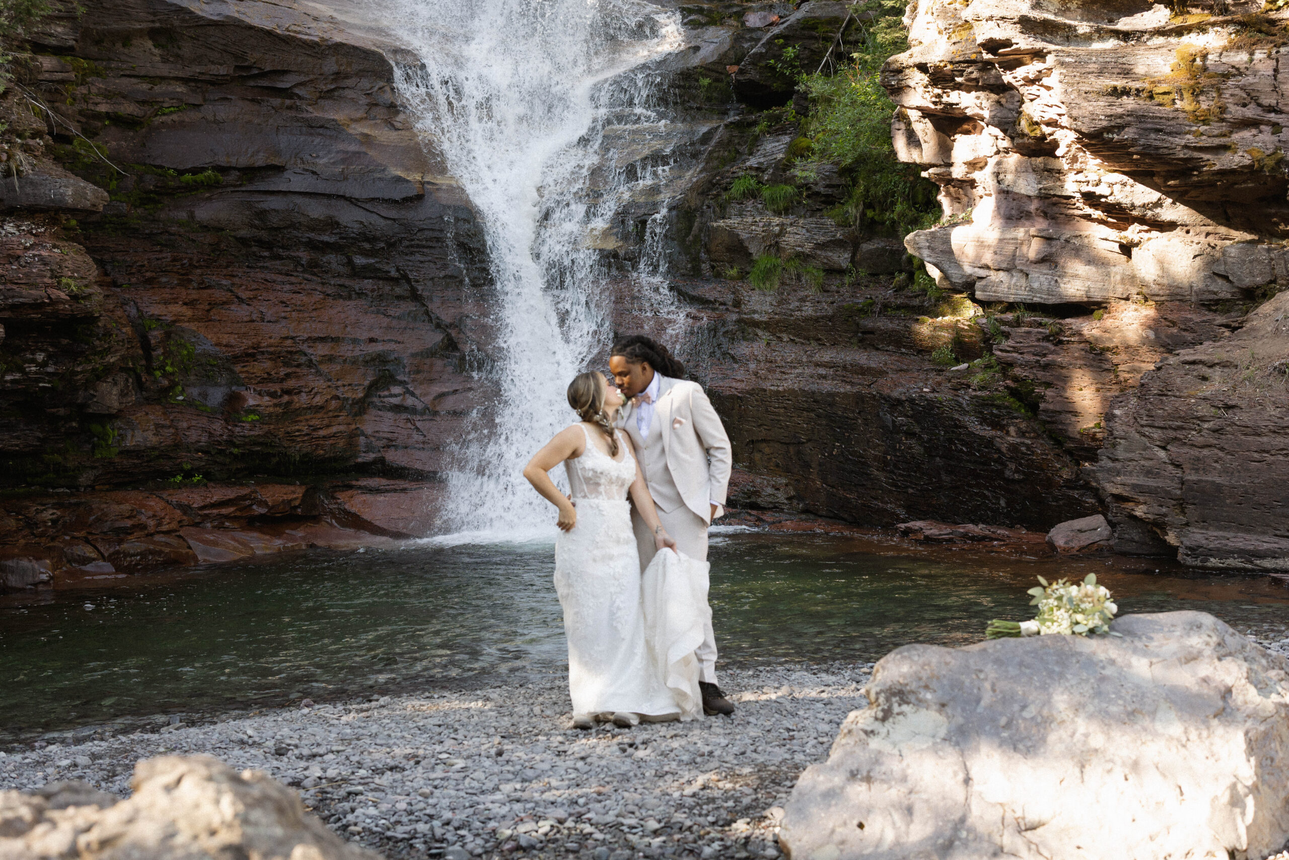 The couple sharing a kiss in front of a waterfall, standing on smooth stones near the water’s edge.
