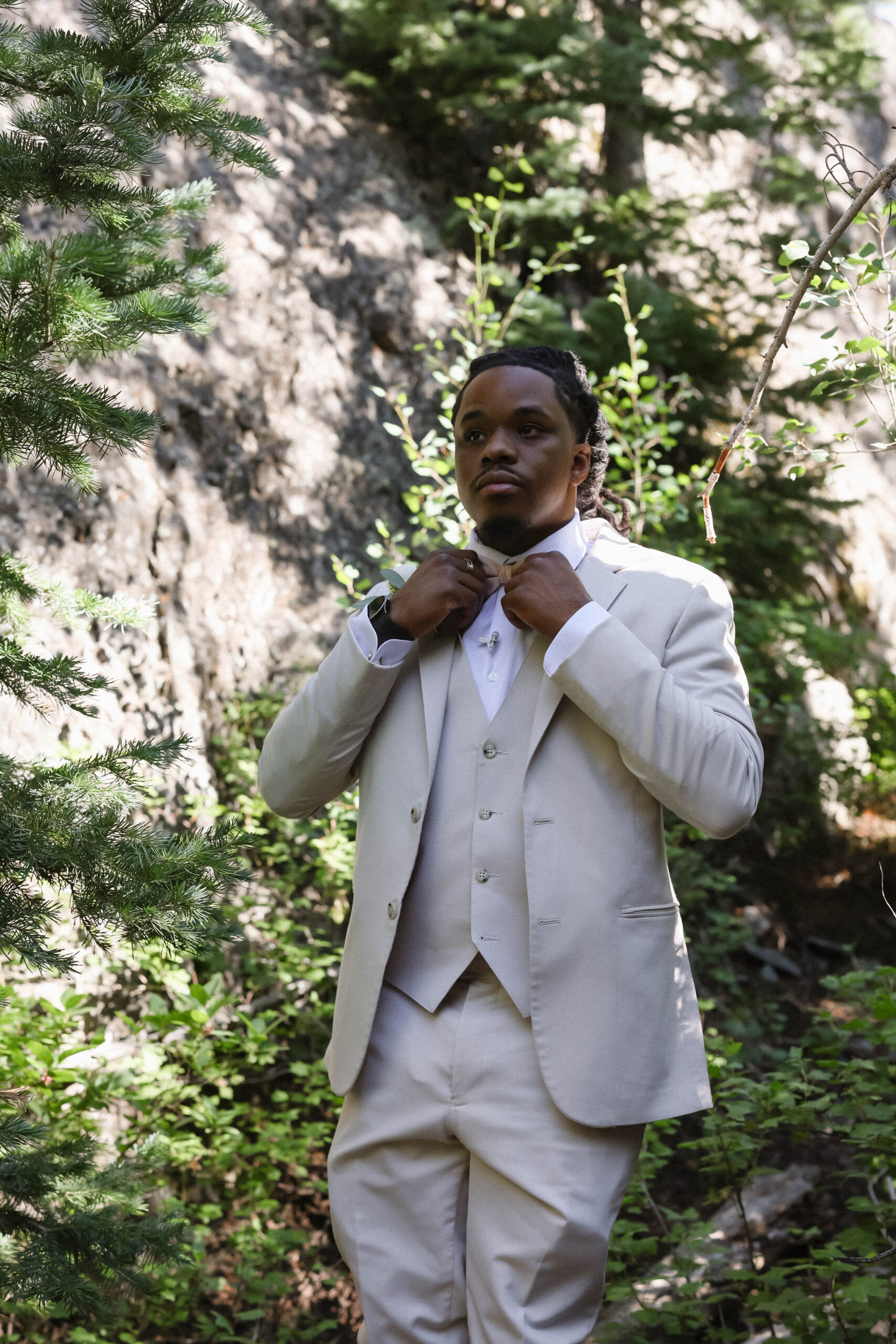 A groom adjusting his bow tie while standing in a shaded forest clearing with sunlight filtering through the trees.