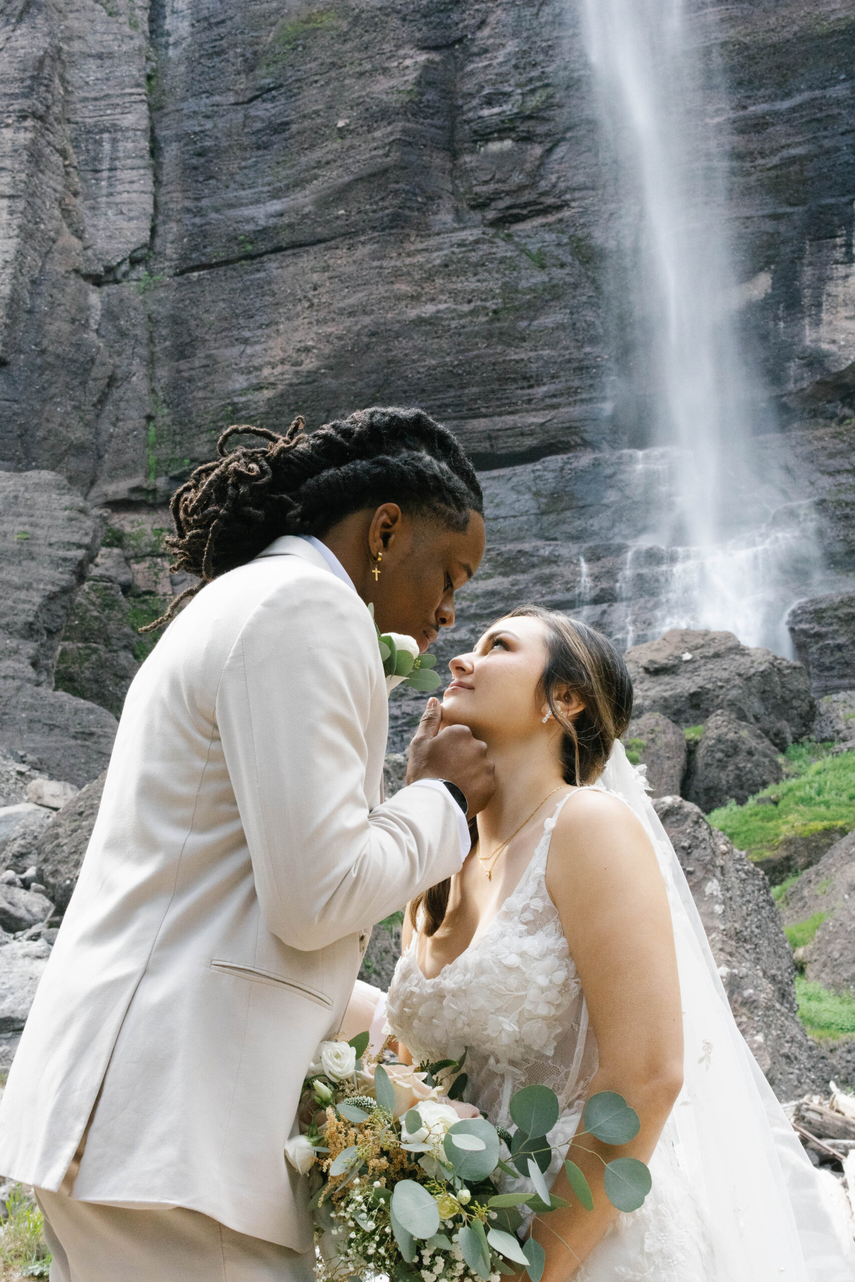An intimate moment at the base of a tall waterfall as the groom gently lifts the bride’s chin to meet his gaze.