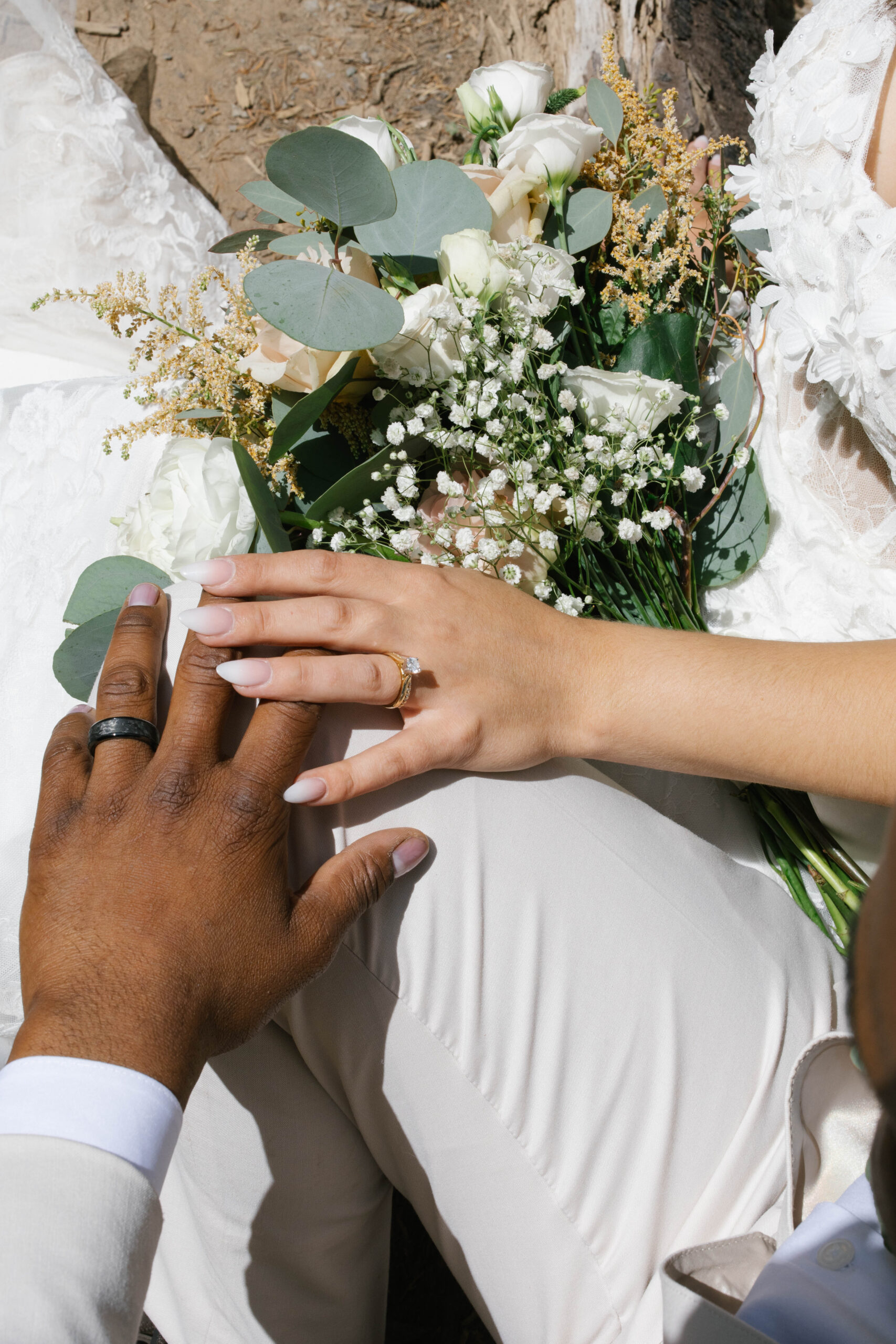 A close-up of the couple’s hands resting together over the bride’s bouquet, showing both wedding rings.