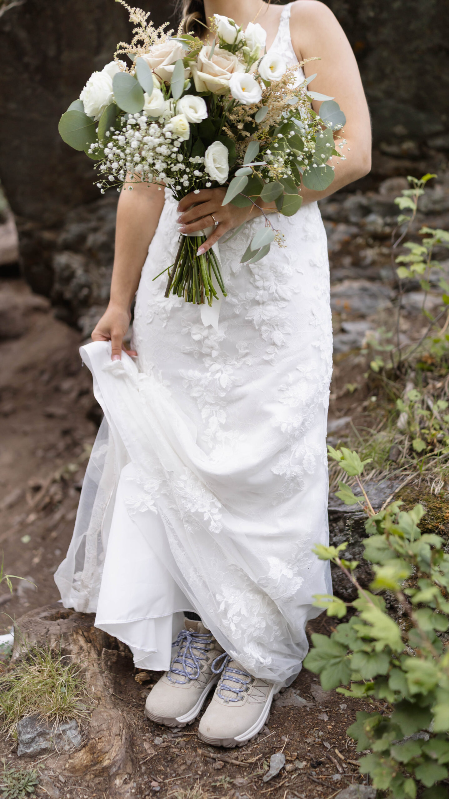 A close-up of the bride’s lace wedding dress paired with hiking boots, with her bouquet held at her waist.