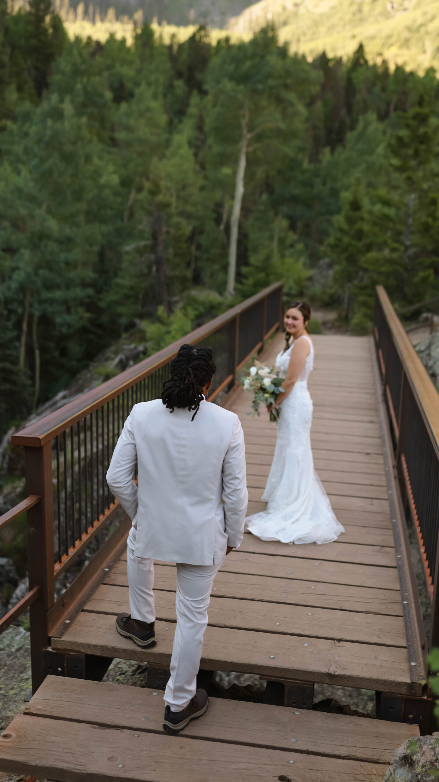 The groom walking up a wooden bridge toward the bride as she looks back over her shoulder.