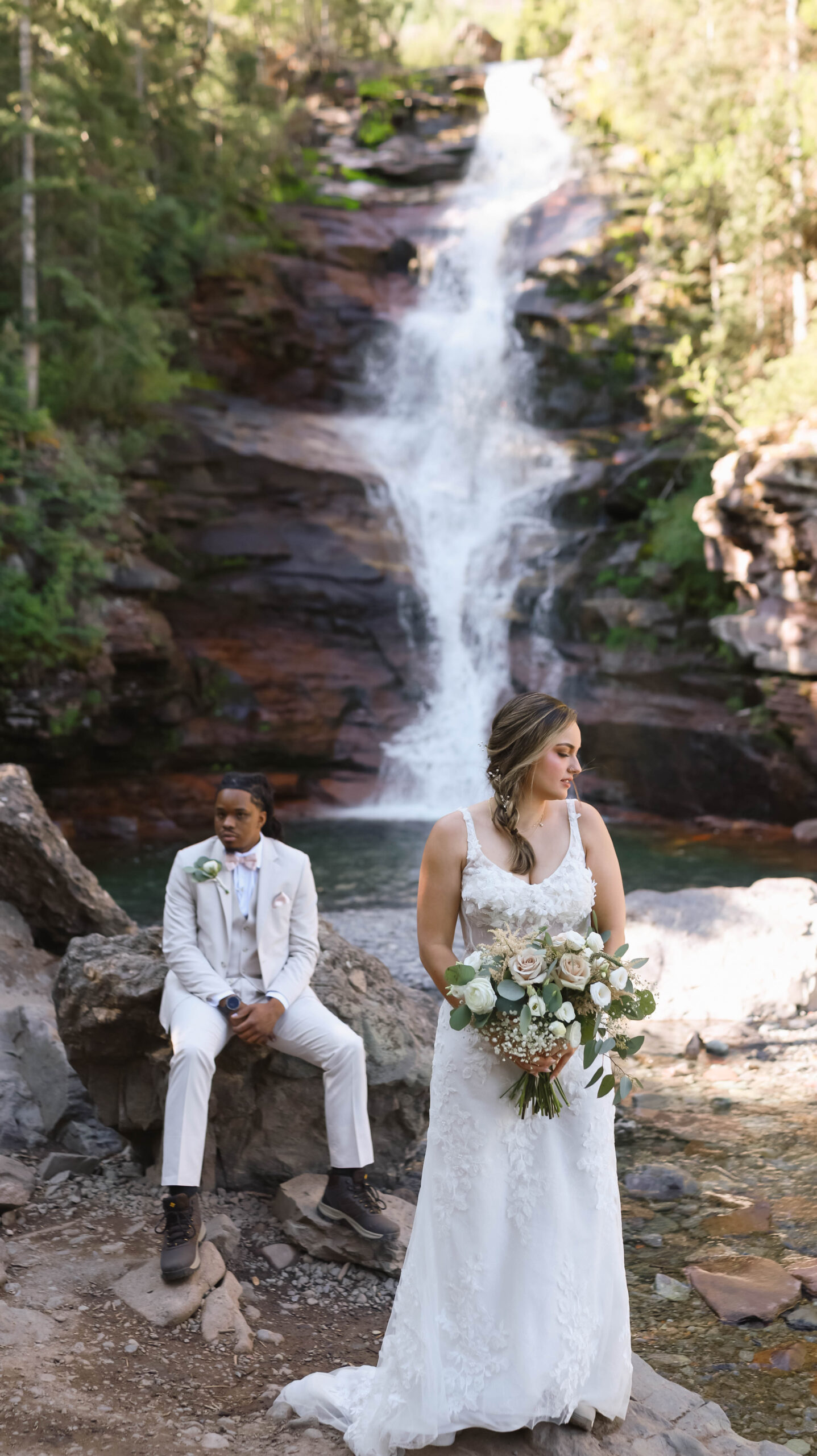 The bride holding her bouquet while the groom sits behind her on a rock, the powerful waterfall cascading into a pool below during their telluride elopement.