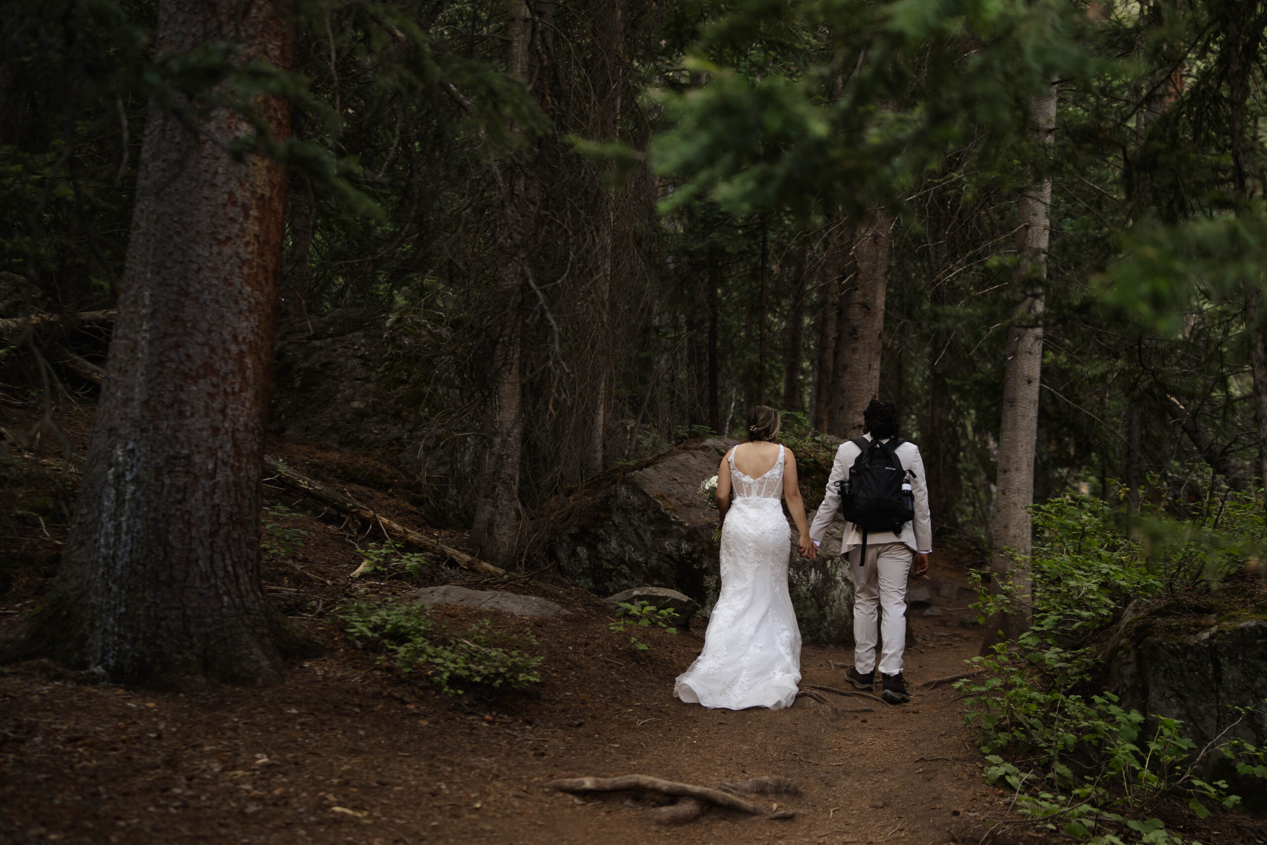 The bride and groom walking hand-in-hand through a dense forest, their wedding attire contrasting with the earthy woodland trail.