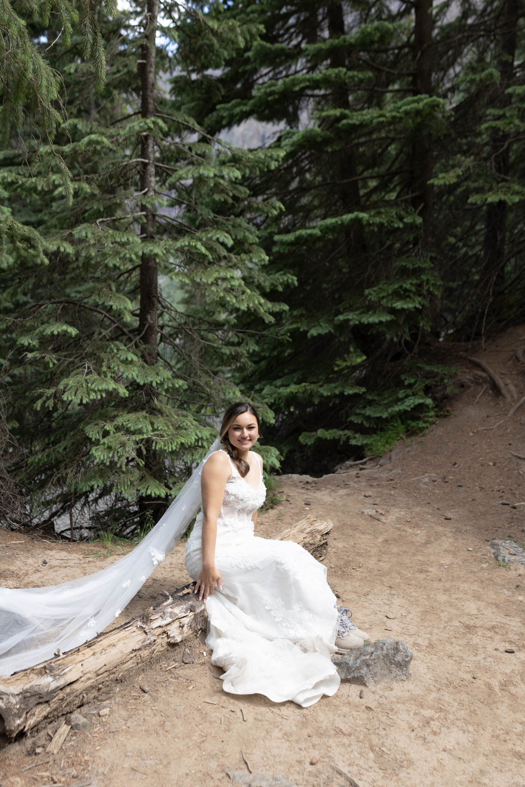 The bride sitting on a fallen tree trunk in the forest, smiling as her dress drapes over the log.