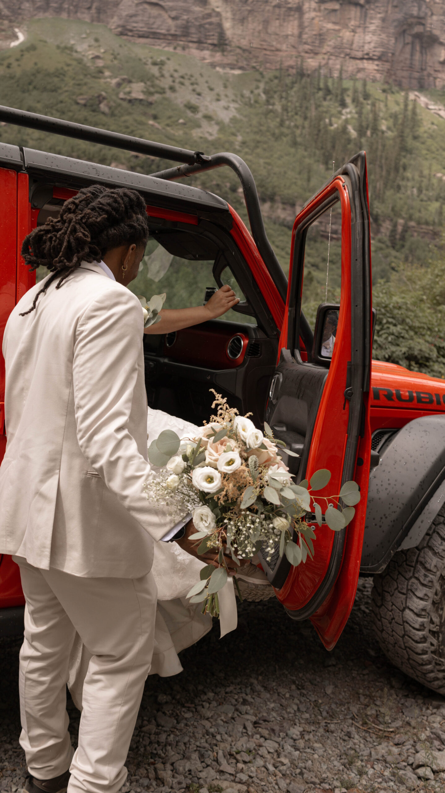 The groom helping the bride into a bright red Jeep, her bouquet overflowing with soft roses and eucalyptus.