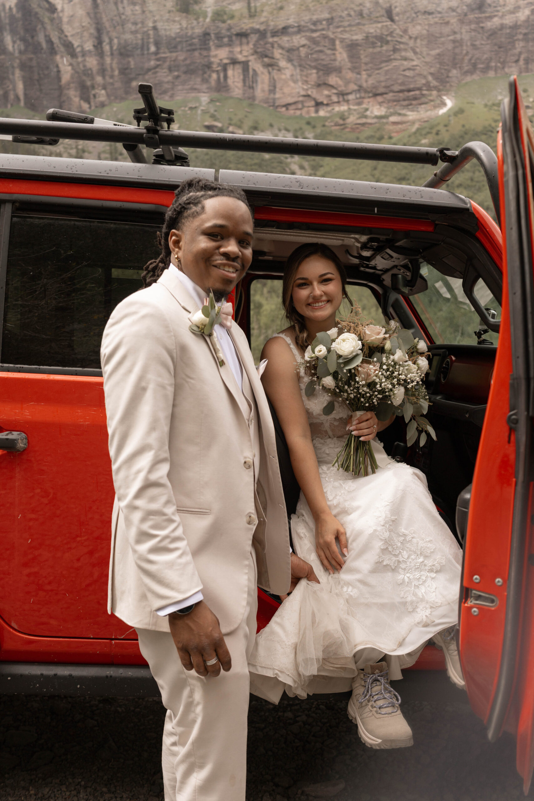 A smiling couple posing beside their red Jeep, the bride holding her bouquet as rocky mountain cliffs rise behind them during their telluride elopement.