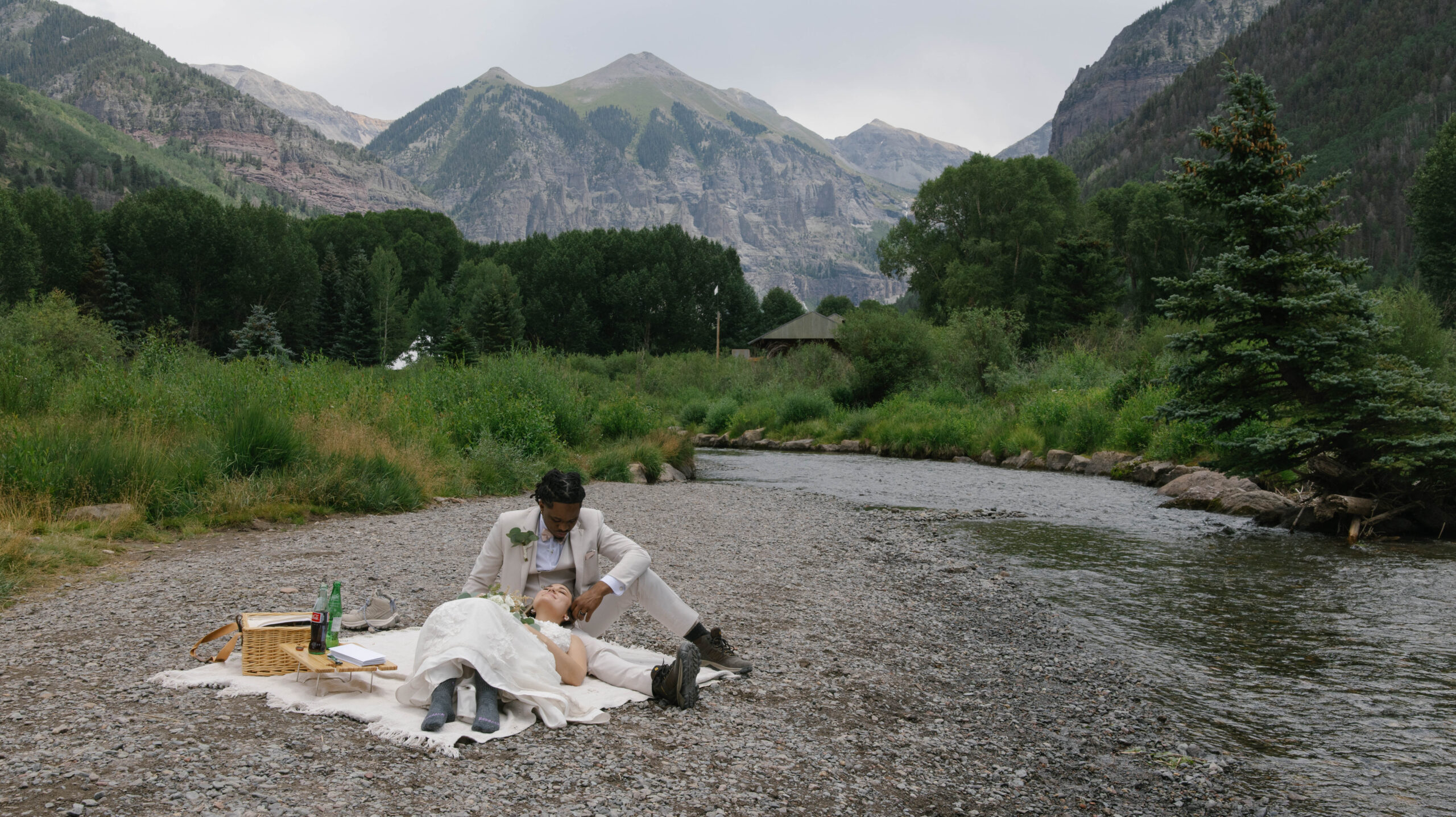 A couple relaxing on a blanket beside a quiet river, surrounded by lush greenery and towering mountains during their telluride elopement.
