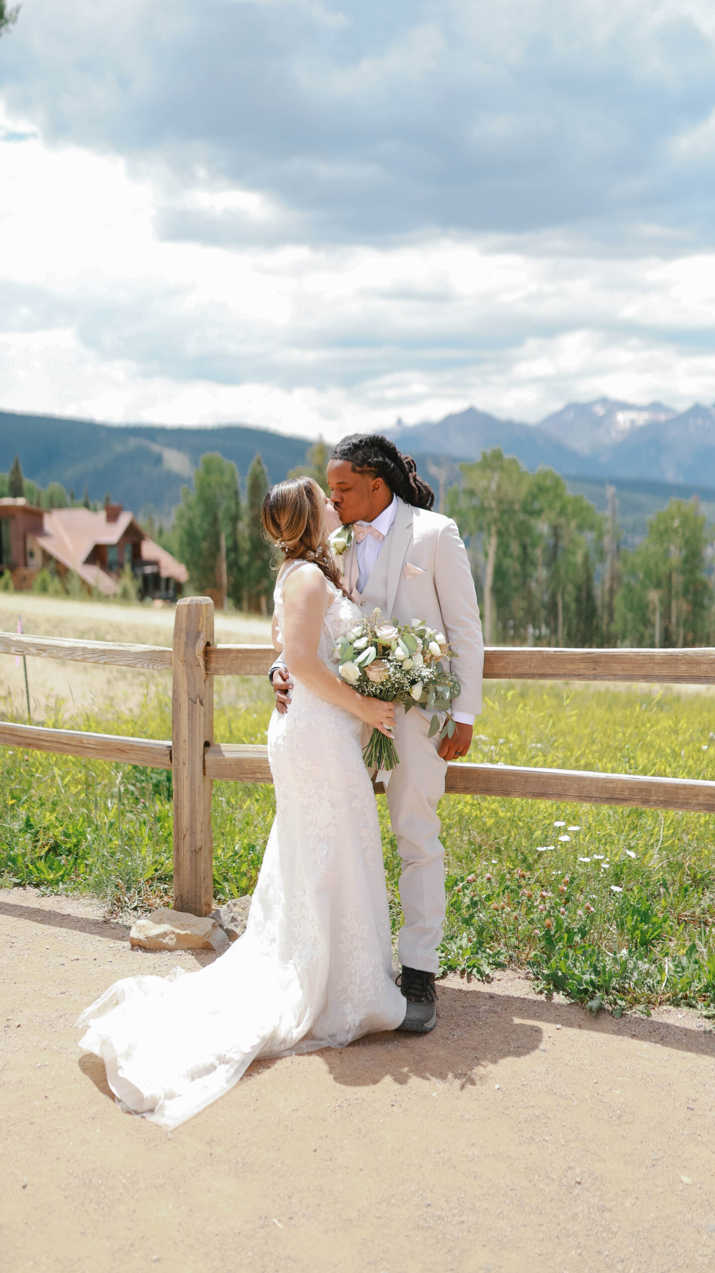 The couple sharing a kiss in front of a rustic wooden fence, surrounded by wildflowers and sweeping mountain views.