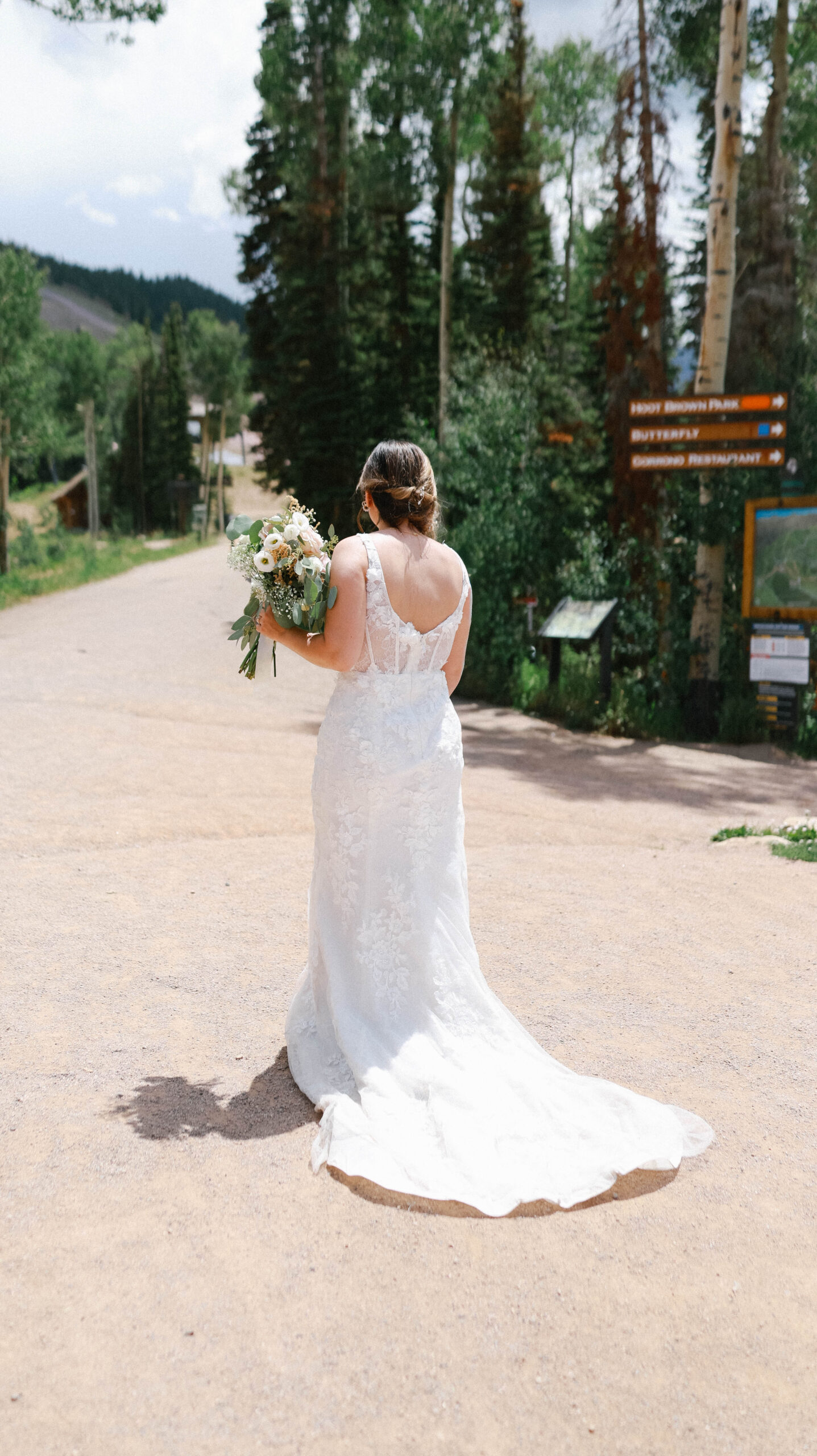 A bride walking alone down a quiet mountain path, her lace gown trailing behind her as she holds her bouquet.