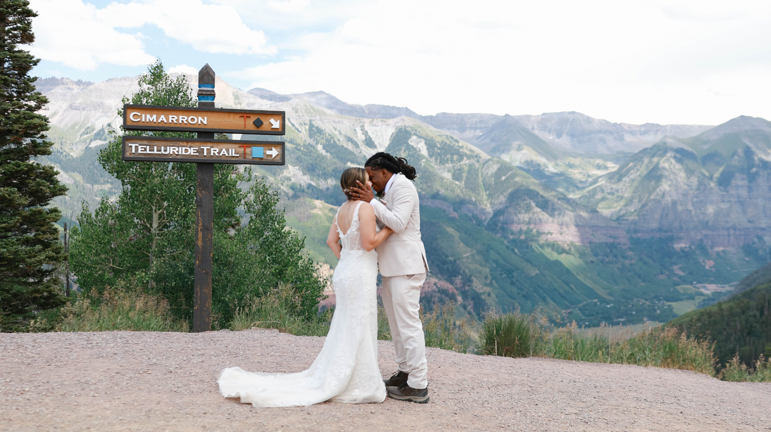 A couple sharing a kiss beneath ski trail signs with expansive mountain ridges unfolding behind them.