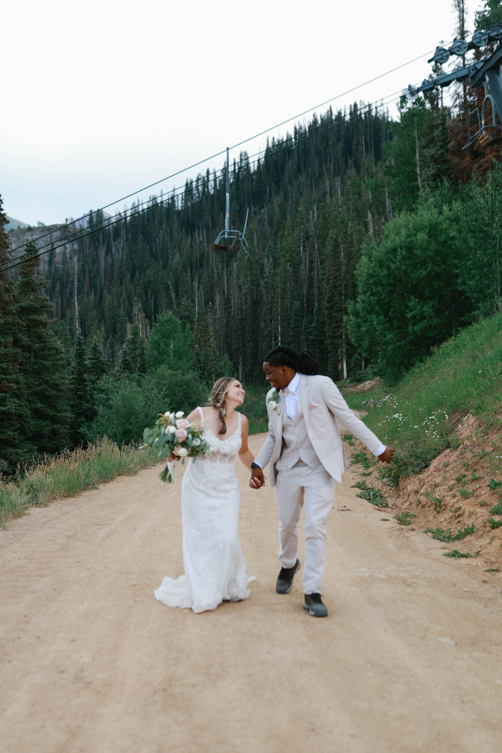 The couple laughing together while walking up a dirt road lined with evergreens and ski-lift cables overhead.