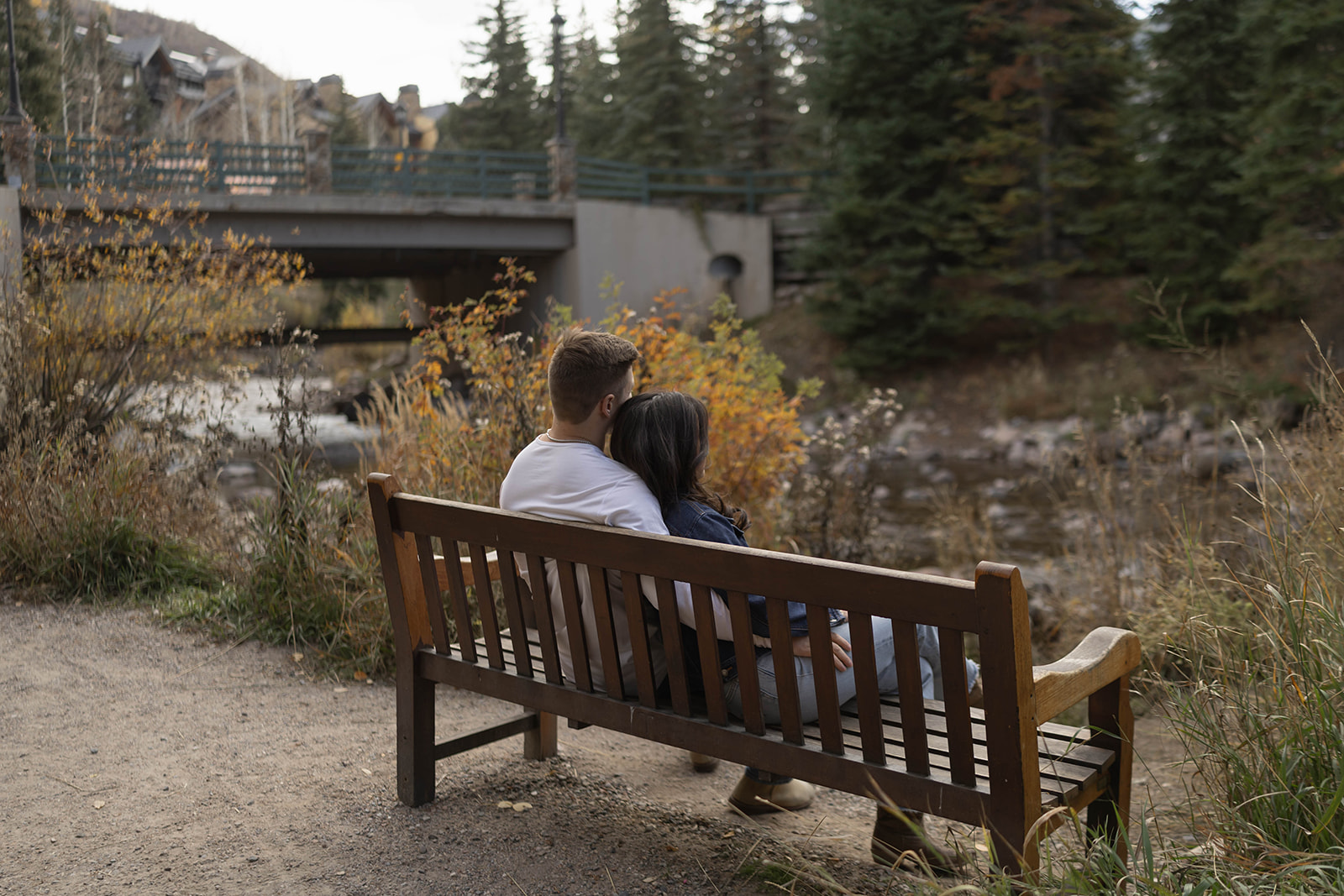 A couple sitting together on a wooden bench beside a flowing creek, framed by autumn foliage and mountain architecture.