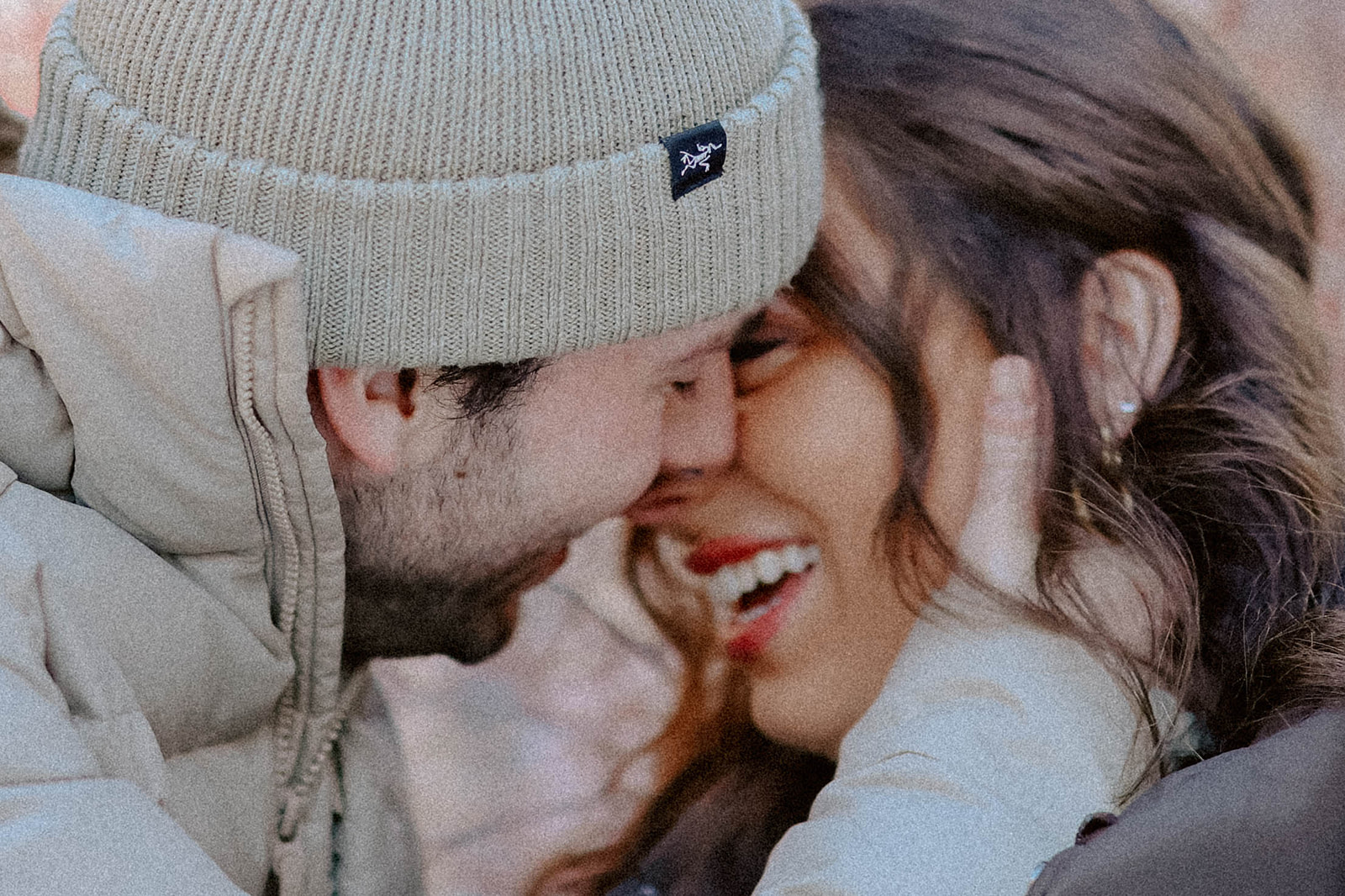 Close-up engagement photo of a couple sharing a joyful kiss while bundled in winter coats and knit hats.