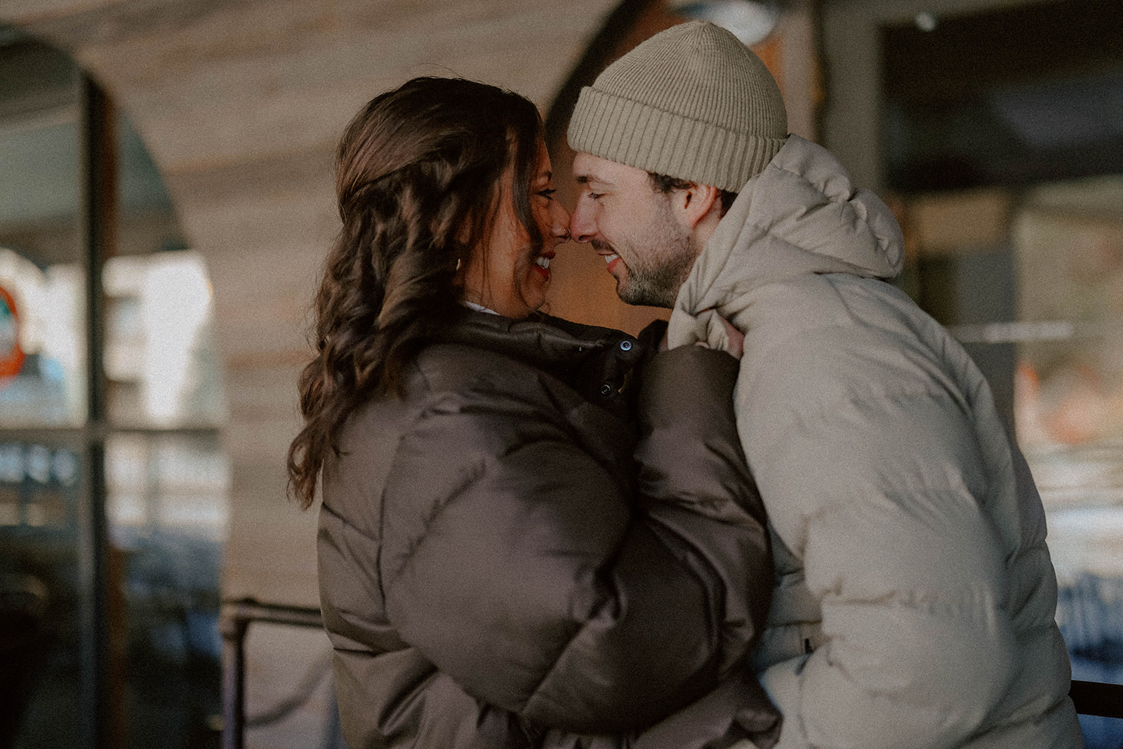 Close-up of a couple laughing and leaning into each other while bundled in cozy winter jackets during a romantic mountain engagement session.