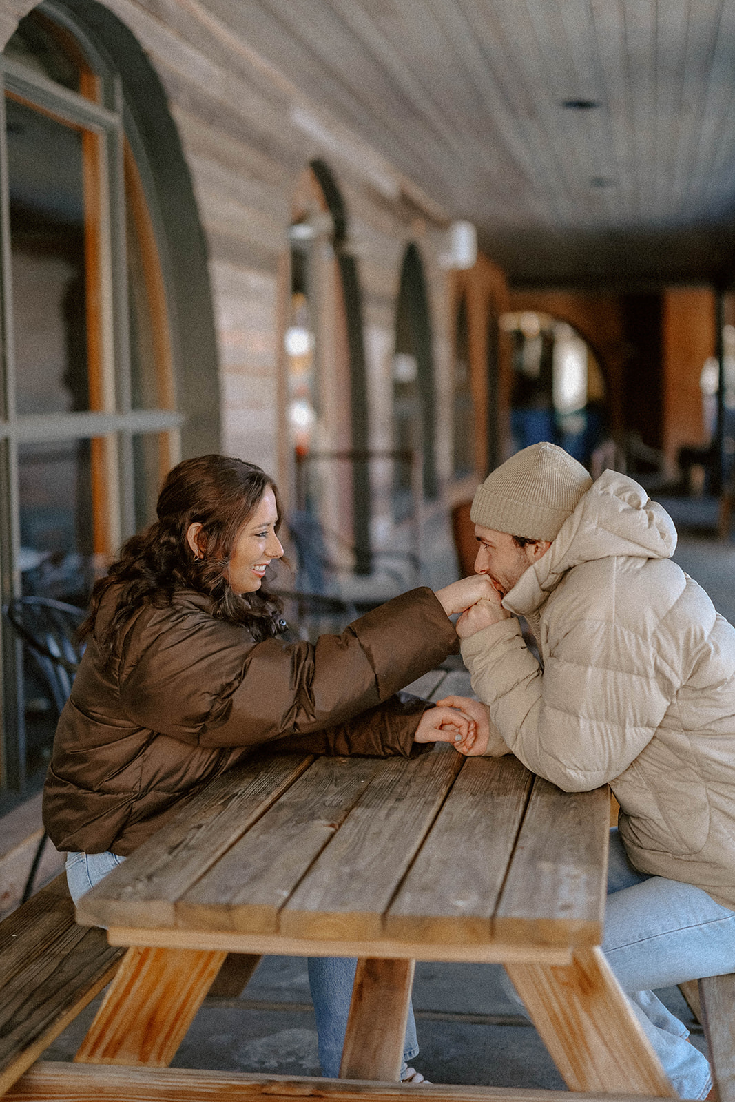 A couple sitting across from each other at a wooden picnic table, bundled in winter layers, sharing a quiet moment in an outdoor café setting.