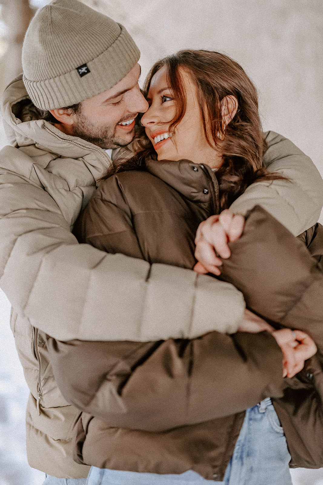 Cozy winter engagement portrait of a couple bundled in puffer jackets, wrapped in each other’s arms and smiling in falling snow.
