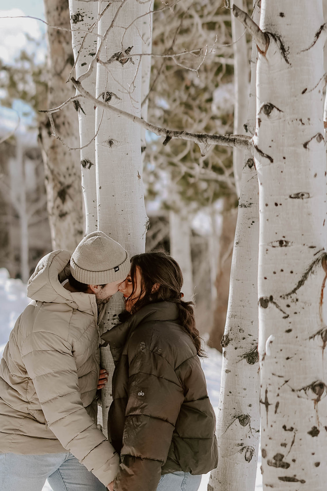 Close-up engagement portrait of a couple touching foreheads and smiling together against a soft evergreen forest background.