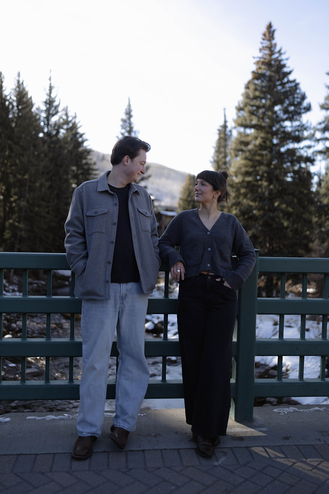A couple leaning casually against a bridge railing, chatting and smiling with mountain views and pine trees in the background in vail village, colorado.