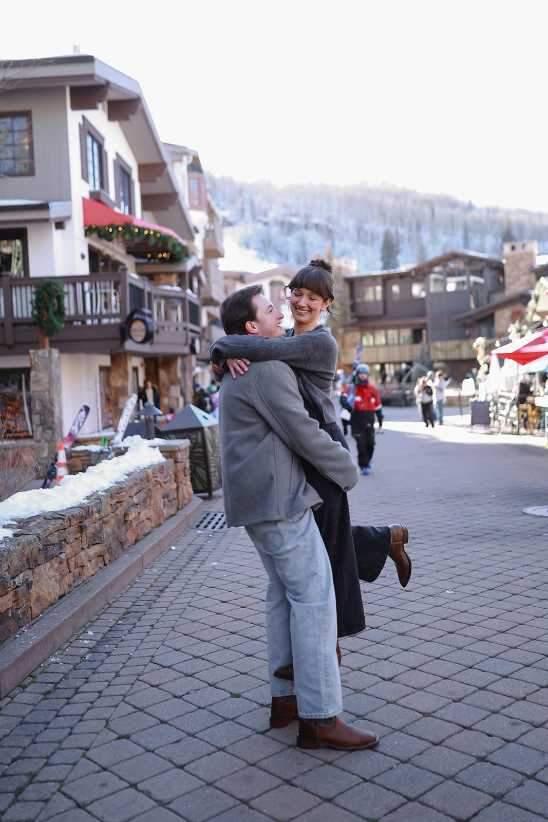 A couple embracing in the middle of a lively pedestrian street with snowy rooftops and cozy shops in the background in vail village, colorado.