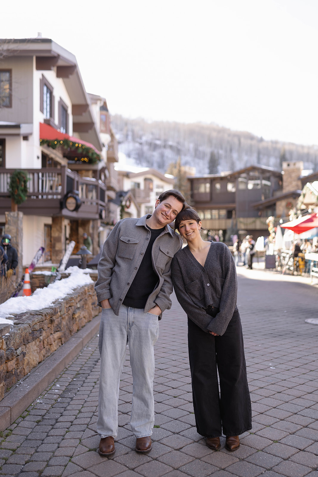 Couple leaning into each other and smiling on a charming alpine street lined with shops and snow.