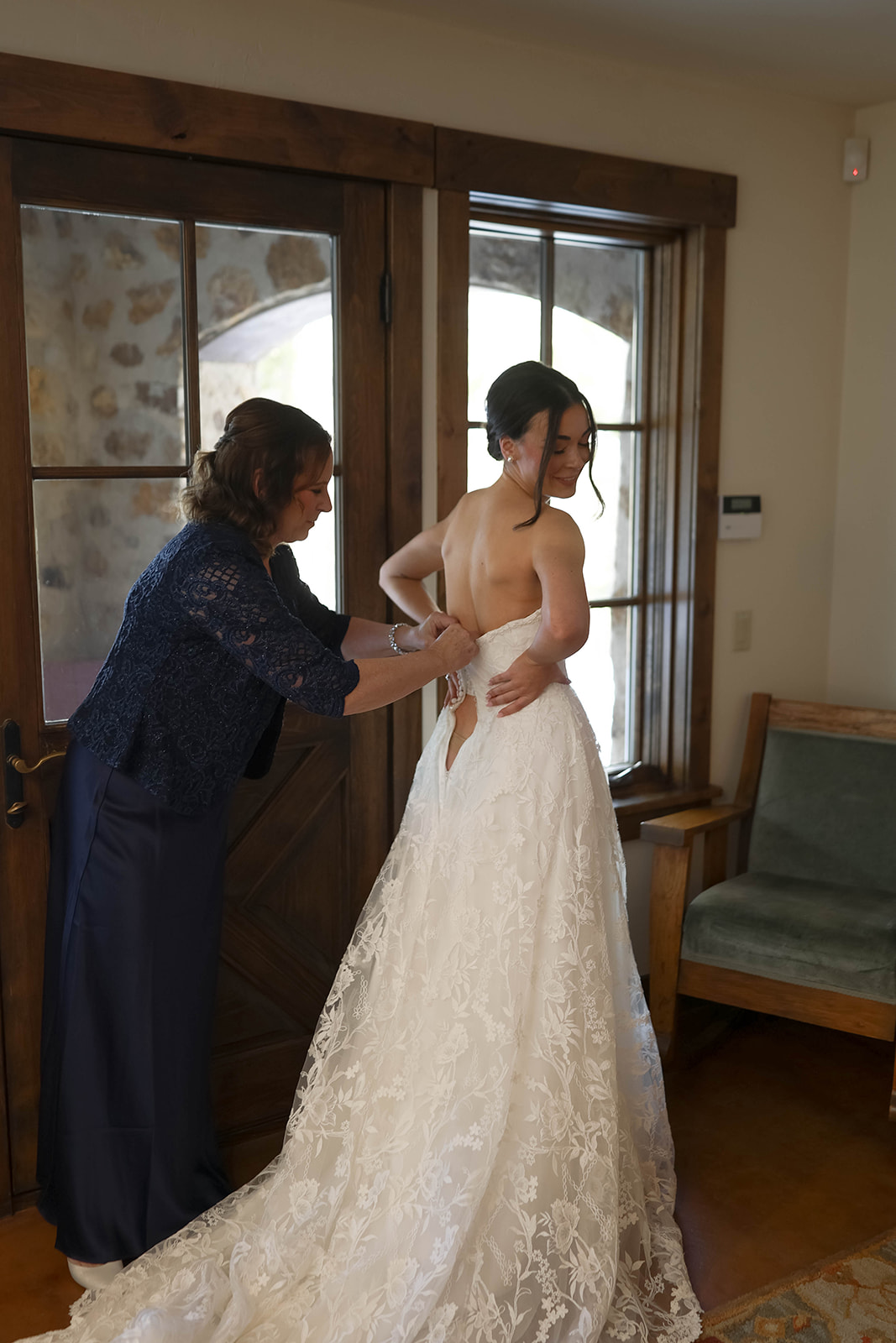 Bride smiling as a family member helps button the back of her lace wedding dress inside a rustic room with wooden trim and stone archway details.