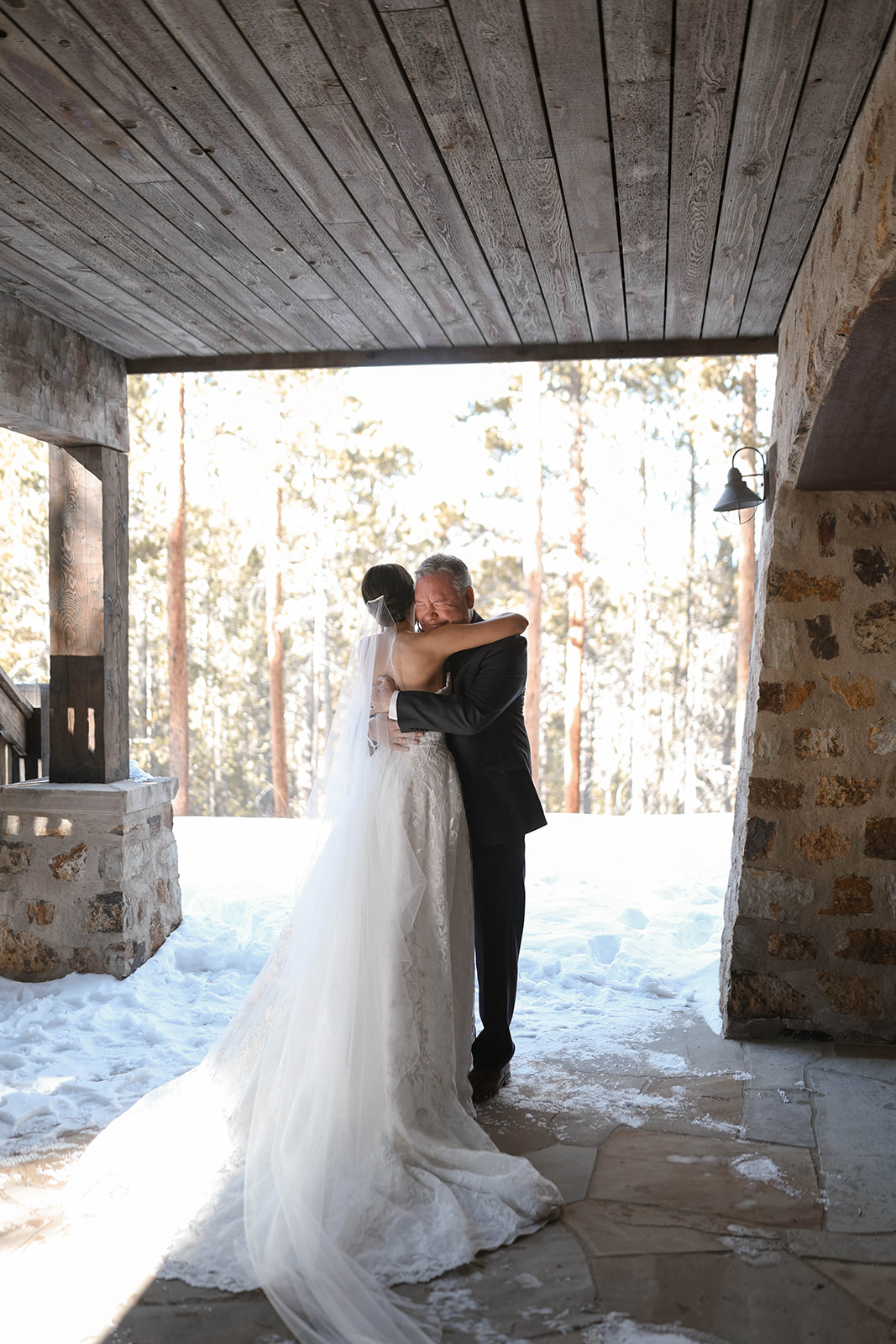 Bride and older man sharing a tight embrace under the porch, her veil trailing behind her in the snow.