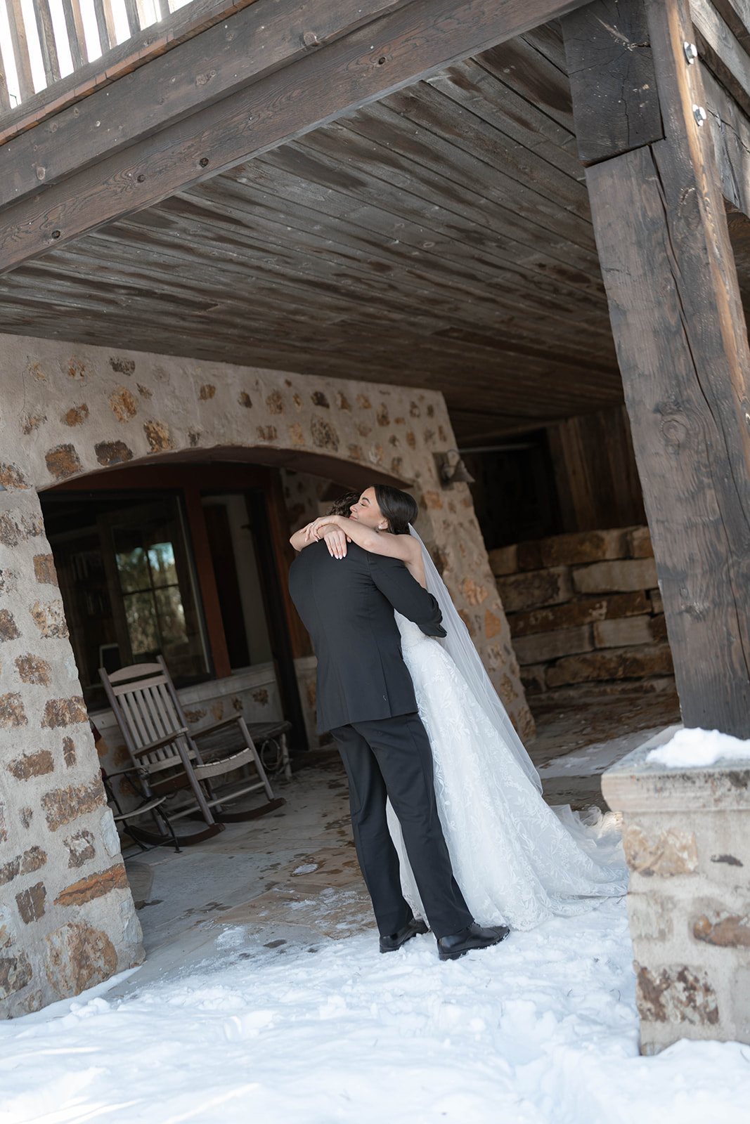 Bride and groom embracing under a rustic wooden porch roof in the snow, the bride’s veil flowing softly behind her.