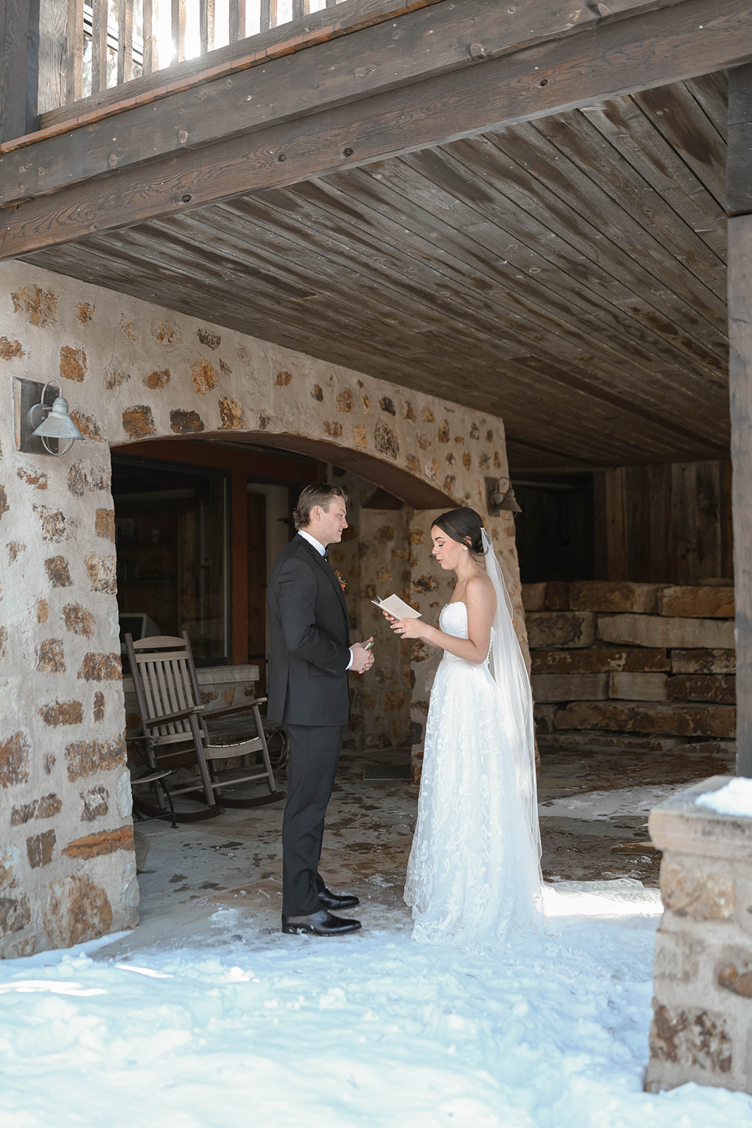 Bride and groom reading their private vows under the wooden covered patio of a stone cabin venue, snow on the ground and ceremony chairs behind them.