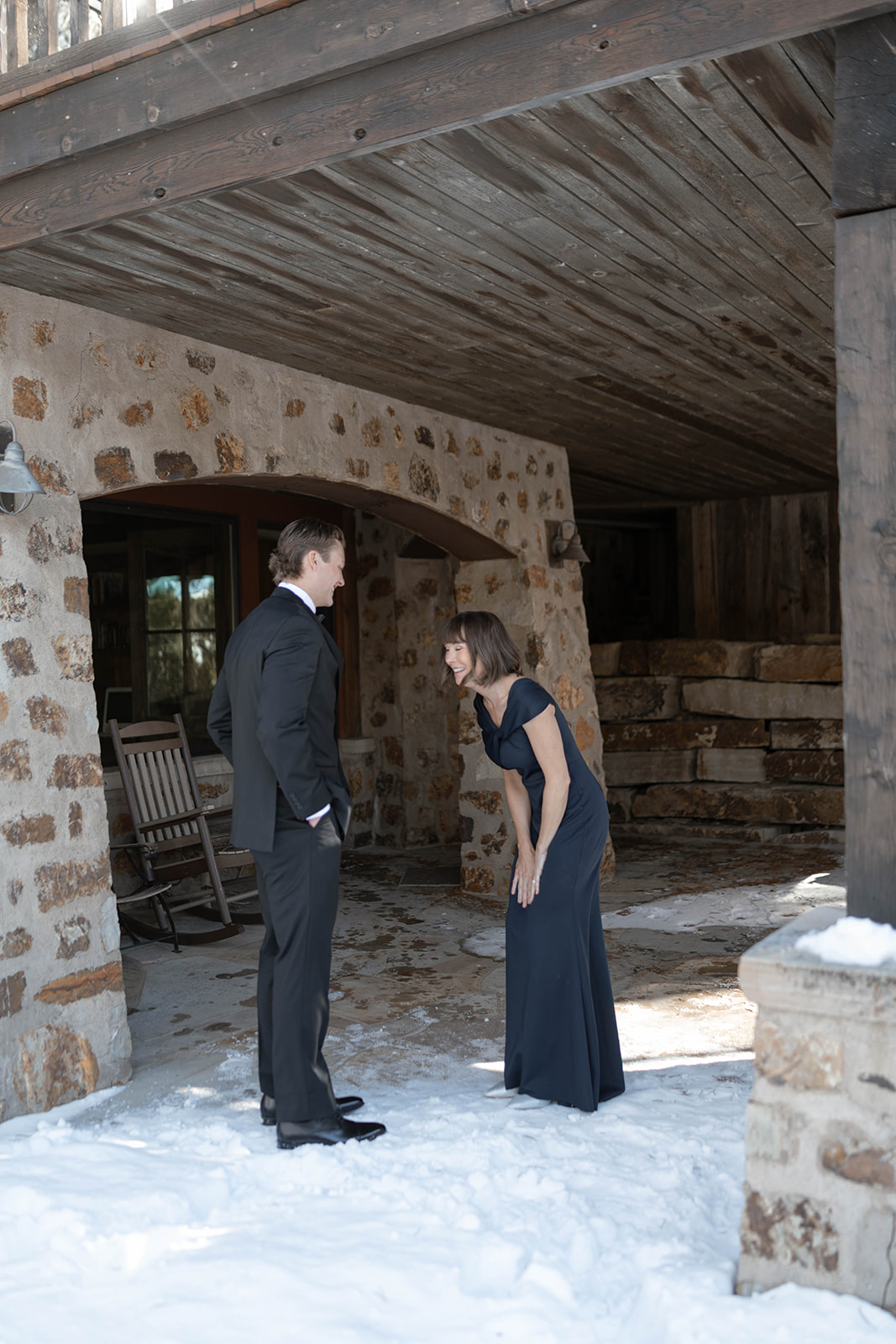 Groom laughing with a woman in a navy dress under a rustic stone and wood porch, snow covering the ground around them.
