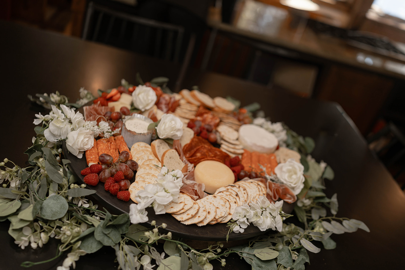 A charcuterie board featuring assorted meats, cheeses, crackers, grapes, and berries, adorned with white flowers and green foliage, creating an elegant and inviting presentation.