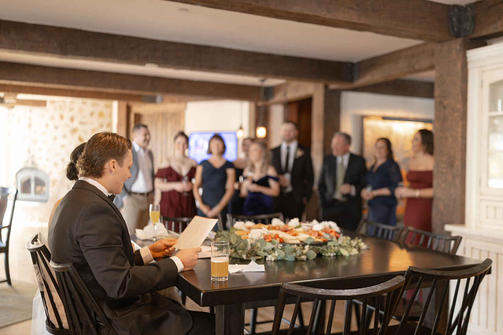 A man in a suit sits at a table, reading a speech, while a group of elegantly dressed people stand in the background, attentively listening.
