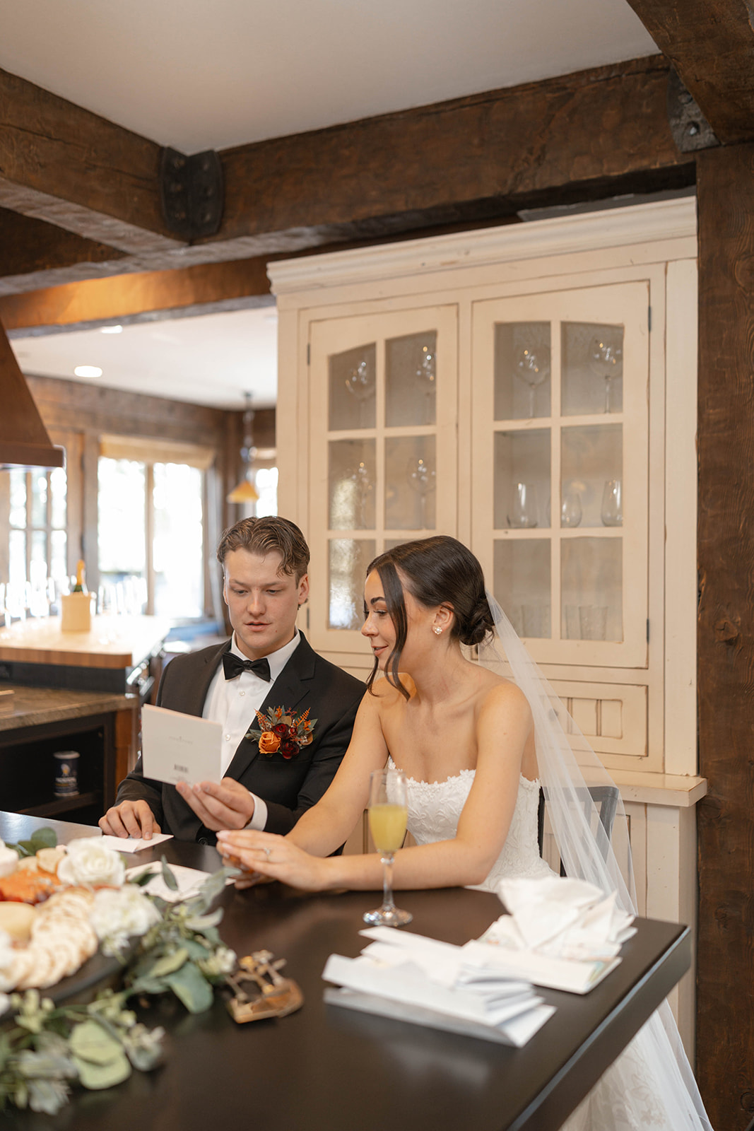 Bride and groom sitting at a wooden table reading cards together inside a cozy cabin-style venue, champagne flute and floral details on the table.