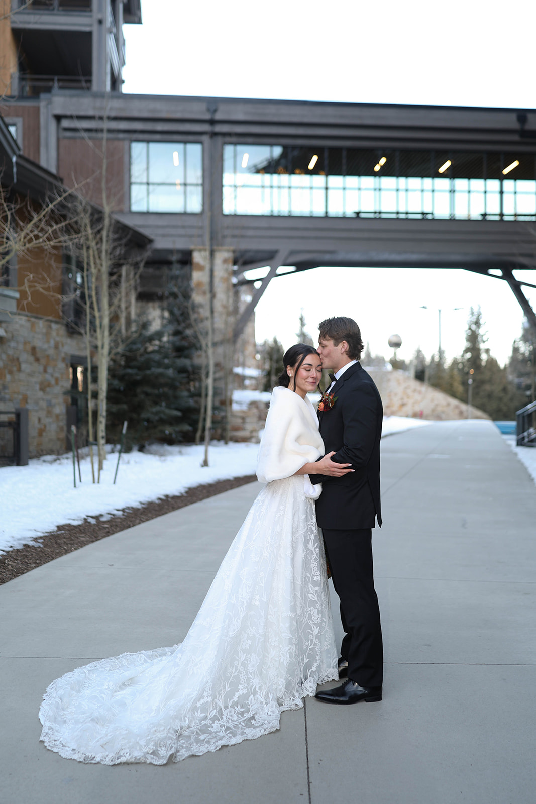 Bride and groom embracing and smiling on a snowy Breckenridge village sidewalk, ski-lodge buildings and a glass skywalk overhead framing the background.