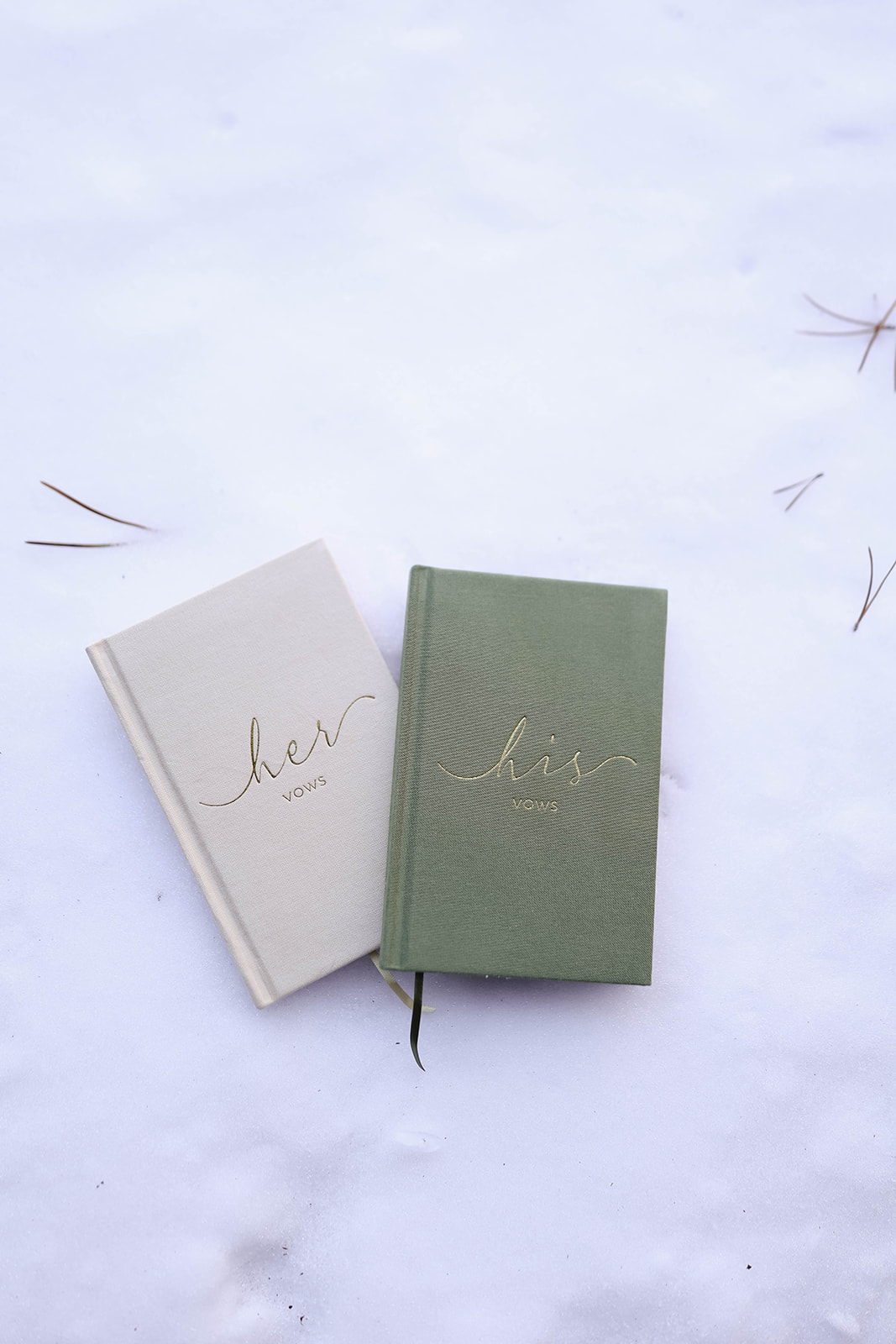 Flat lay of two vow books on snow-covered ground, one cream-colored and one sage green, both with gold embossed “vows” lettering.