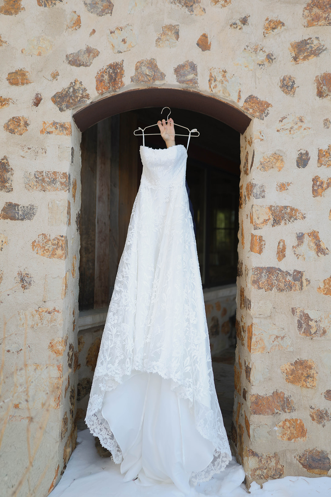 White wedding dress with lace details hangs on a wooden hanger in a rustic stone archway. The scene conveys elegance and anticipation.