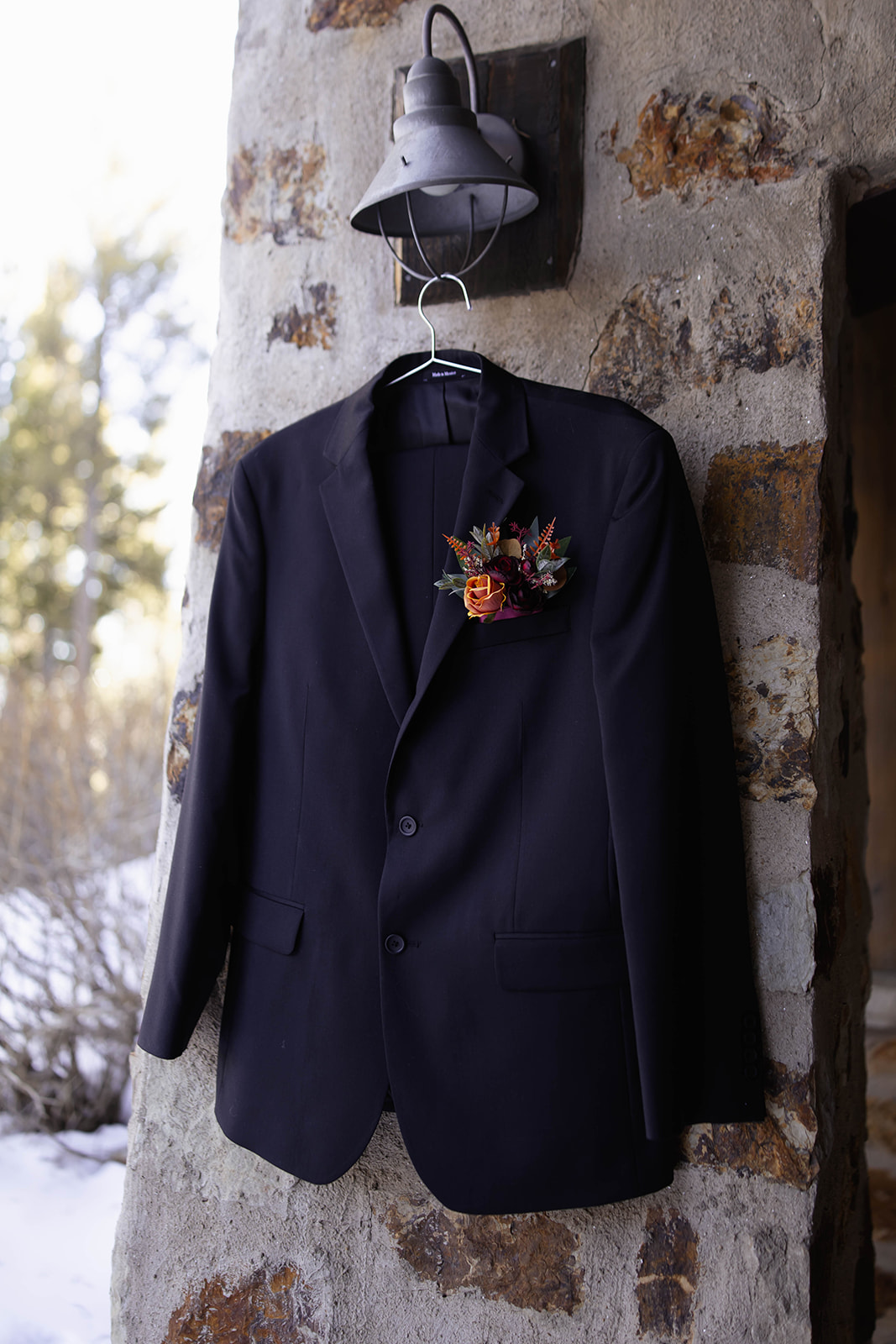 Groom’s black suit hanging on a textured stone wall under a metal lantern, featuring a rose and greenery boutonniere pinned on the lapel.