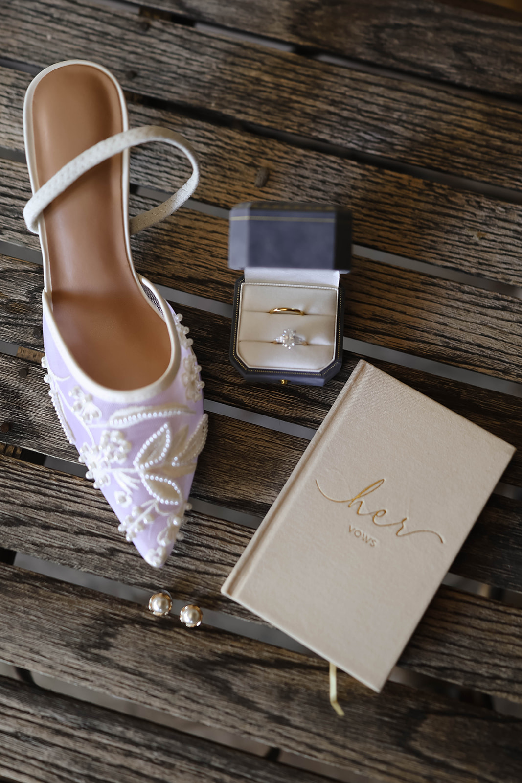White bridal slipper with pearl embellishments, a ring box with two gold rings, a "her vows" book, and pearl earrings on rustic wood. Elegant and romantic.