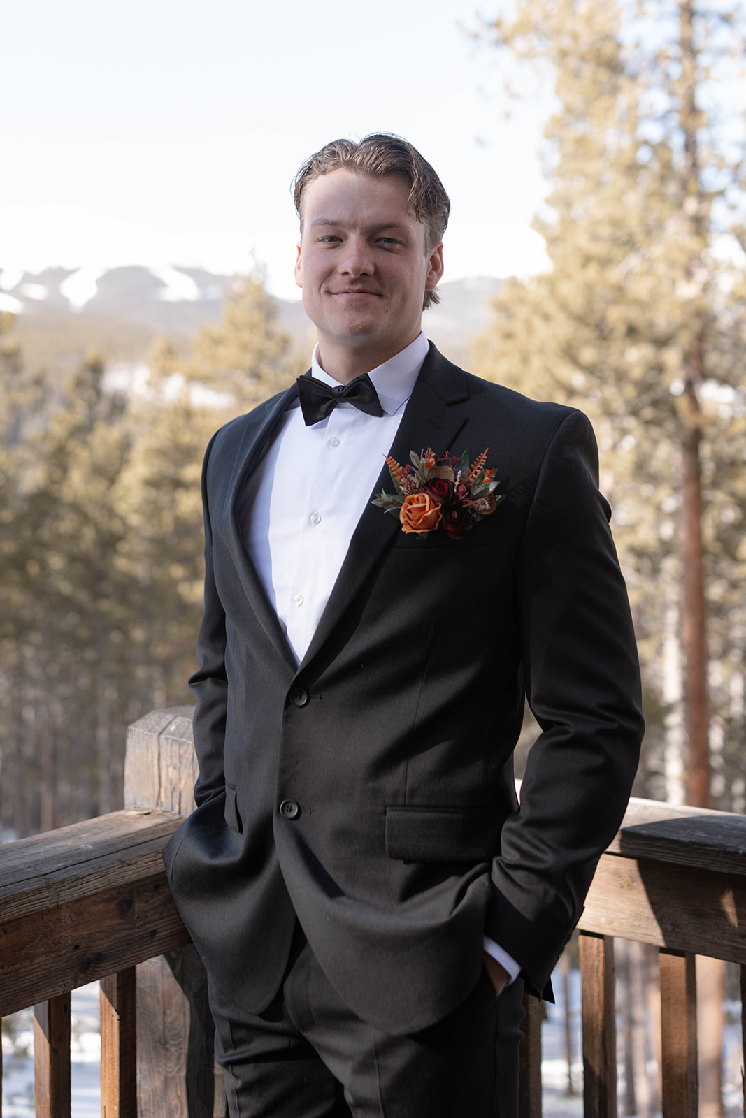 Man in a black suit and bow tie stands outdoors with hands in pockets, ready for his Breckenridge elopement. He has a flower boutonniere and stands against a background of snowy mountains and trees.