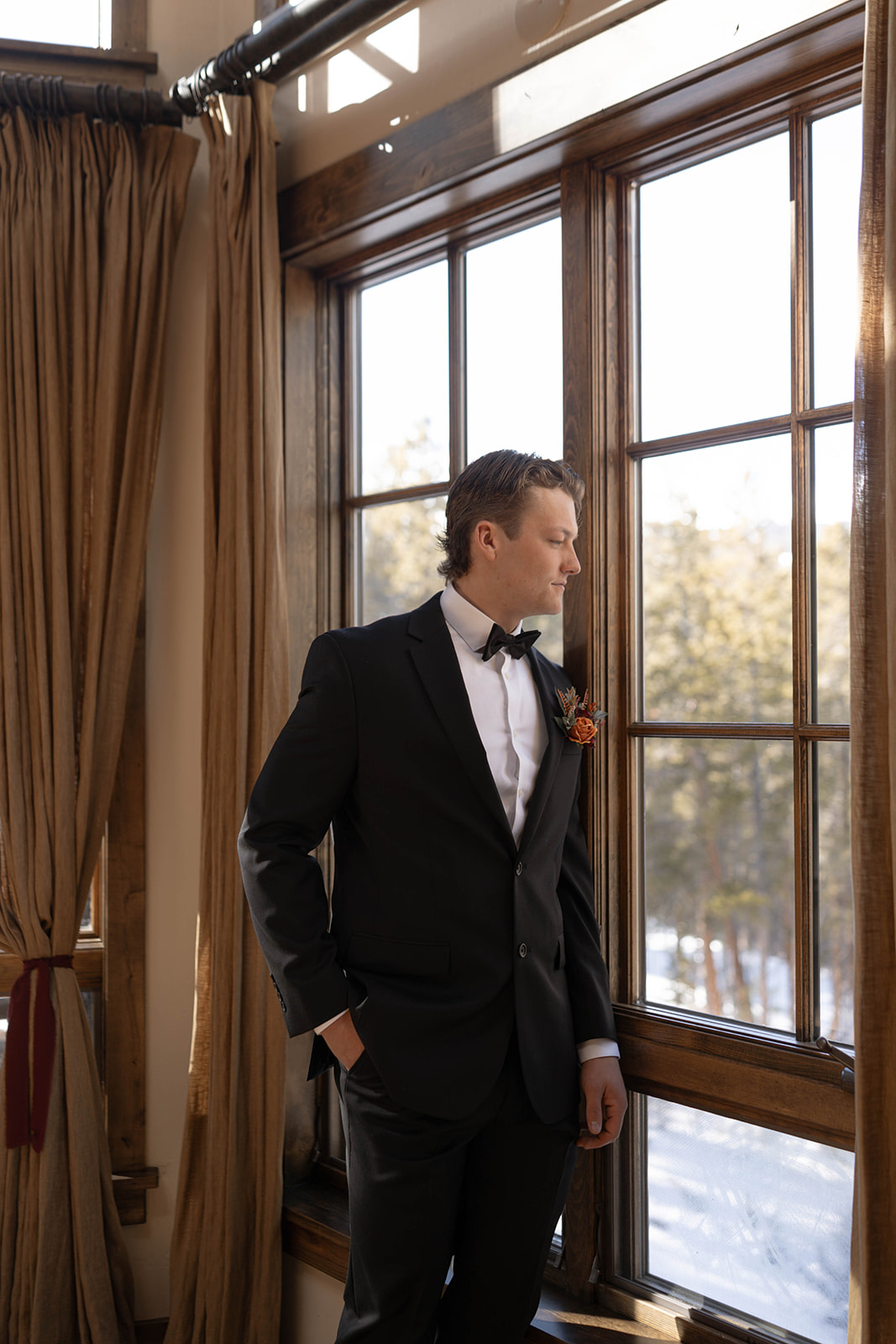 Groom standing by a large window inside a rustic cabin, wearing a black tuxedo with a boutonniere and looking out toward snow-covered trees, captured during a Breckenridge elopement.