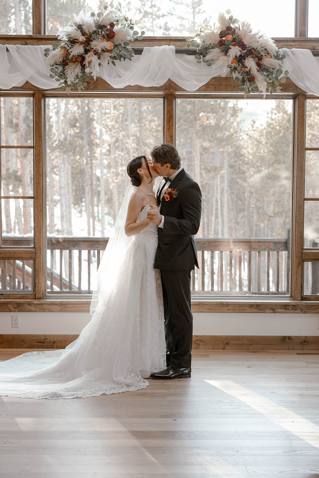A bride and groom share a kiss indoors by large windows with snowy trees outside during their Breckenridge elopement. Elegant floral arrangements adorn the window top, adding romance.