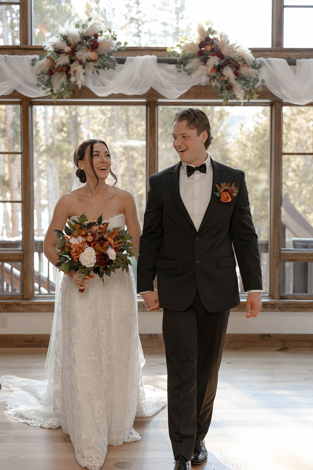 Bride and groom walking hand in hand inside a light-filled mountain venue, smiling at each other beneath large windows decorated with white draping and floral arrangements.
