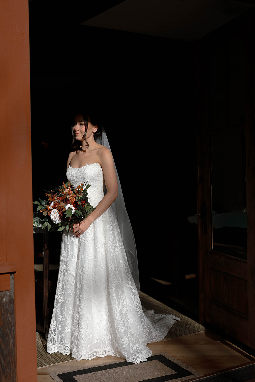 Bride standing alone with her bouquet in warm directional light peeking through a cabin doorway, moody and editorial portrait from their Breckenridge elopement day.