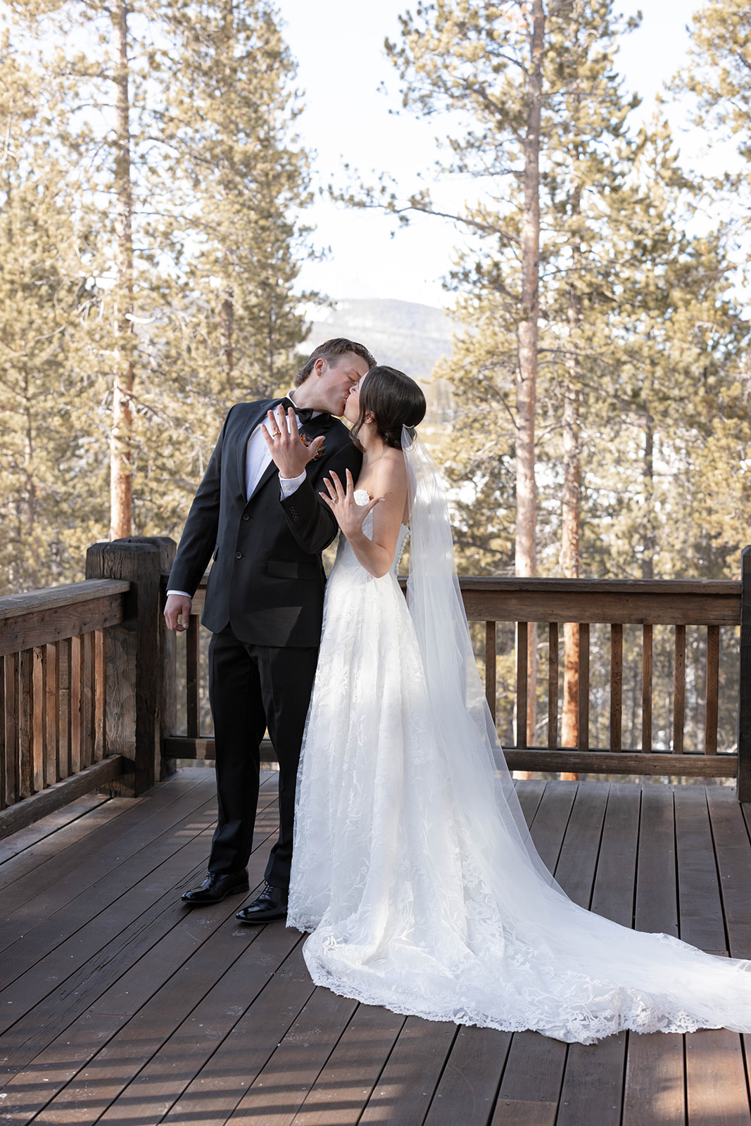 A bride and groom share a kiss on a wooden deck, surrounded by tall pine trees in a serene forest setting. The bride wears a lace gown; the groom is in a suit.