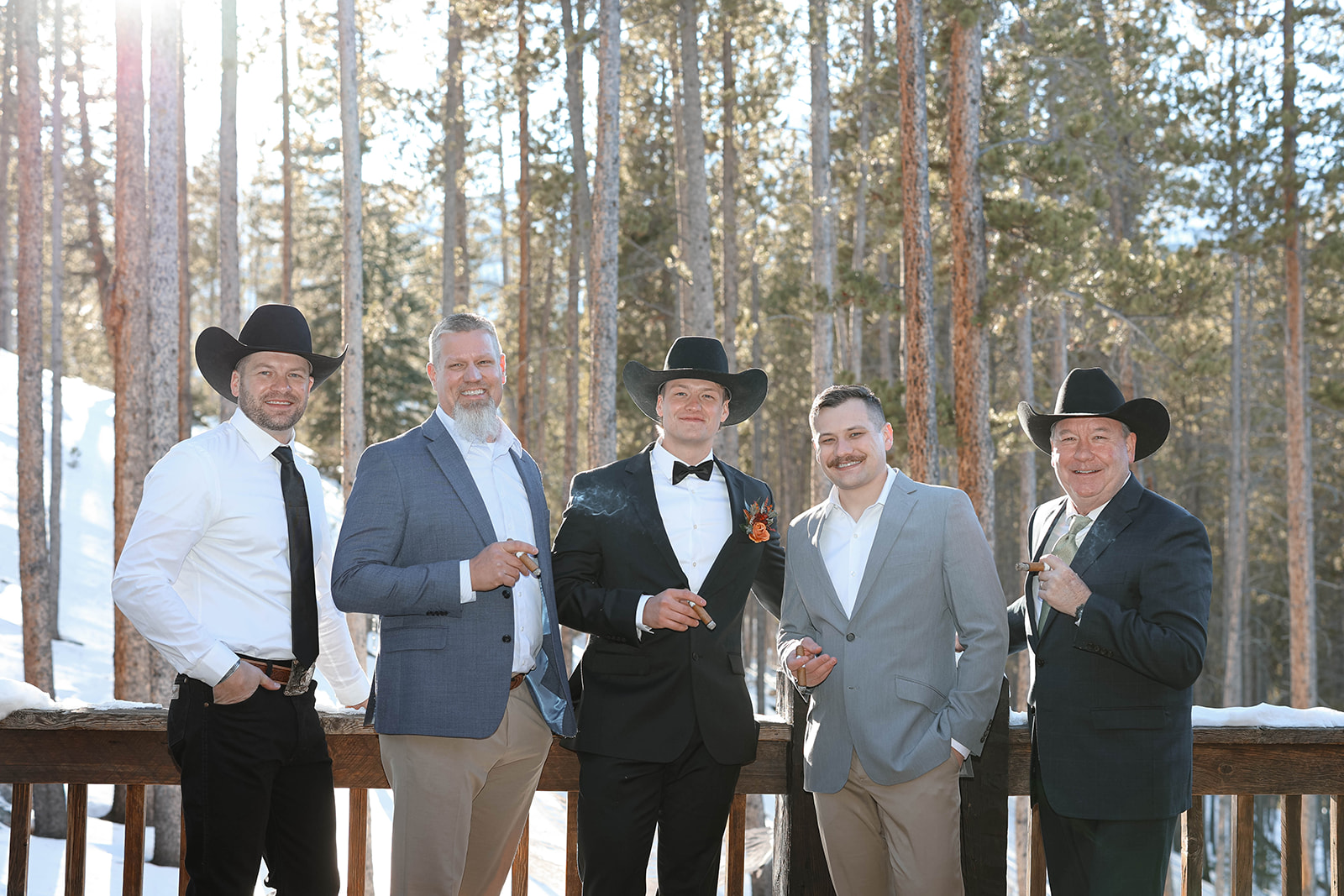 Groom and his groomsmen in suits and cowboy hats holding cigars, standing on a wooden deck surrounded by pine trees during a Breckenridge elopement celebration.