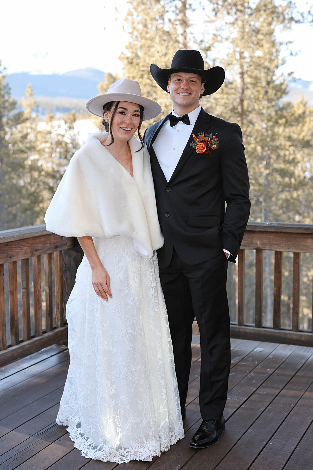 A couple in formal attire smiles on a wooden deck. The woman wears a white dress and hat, the man a black suit with a cowboy hat. Forest backdrop.