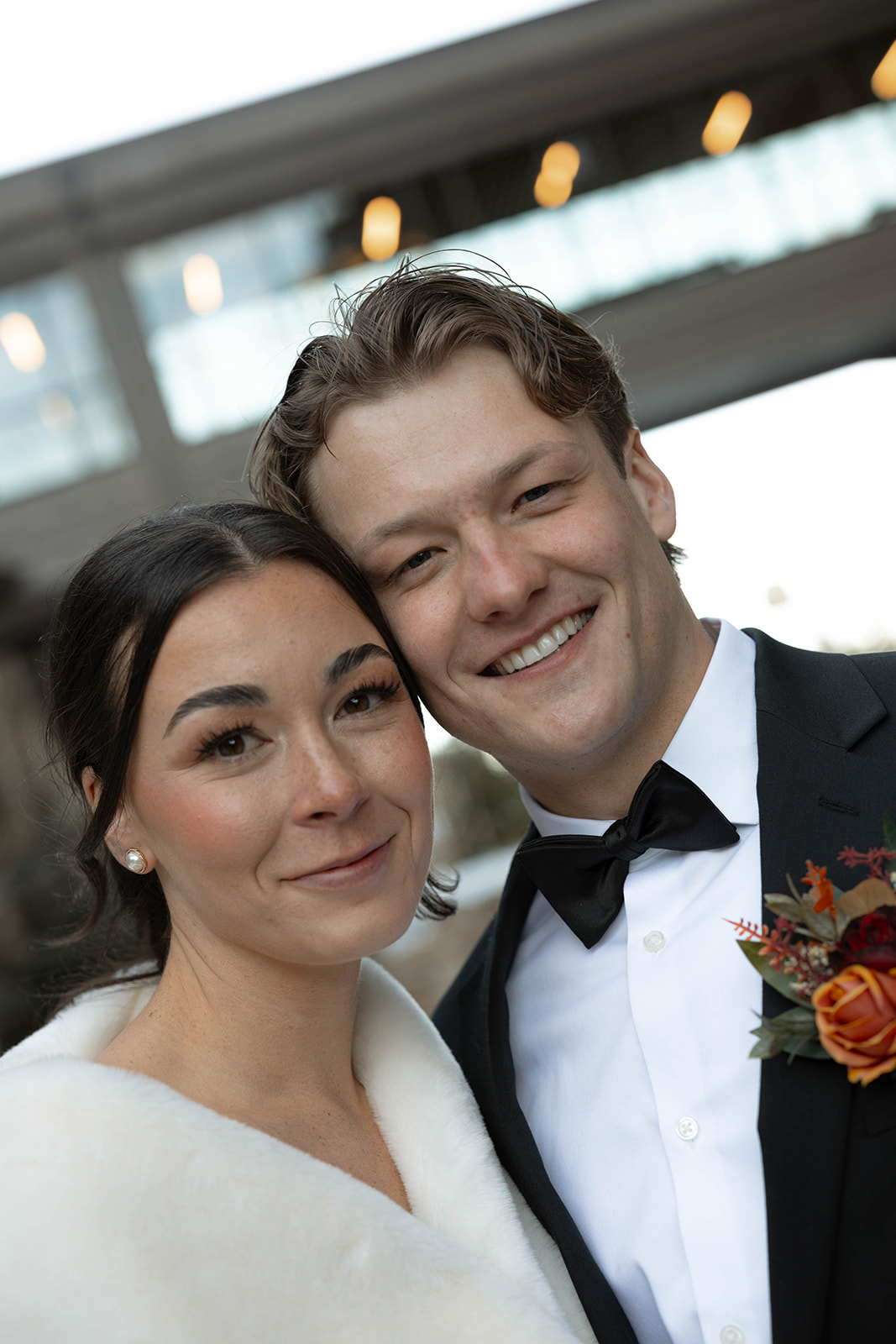 A couple smiles warmly on their wedding day. The woman wears a white dress with a fur shawl, and the man dons a black suit with a floral boutonniere.