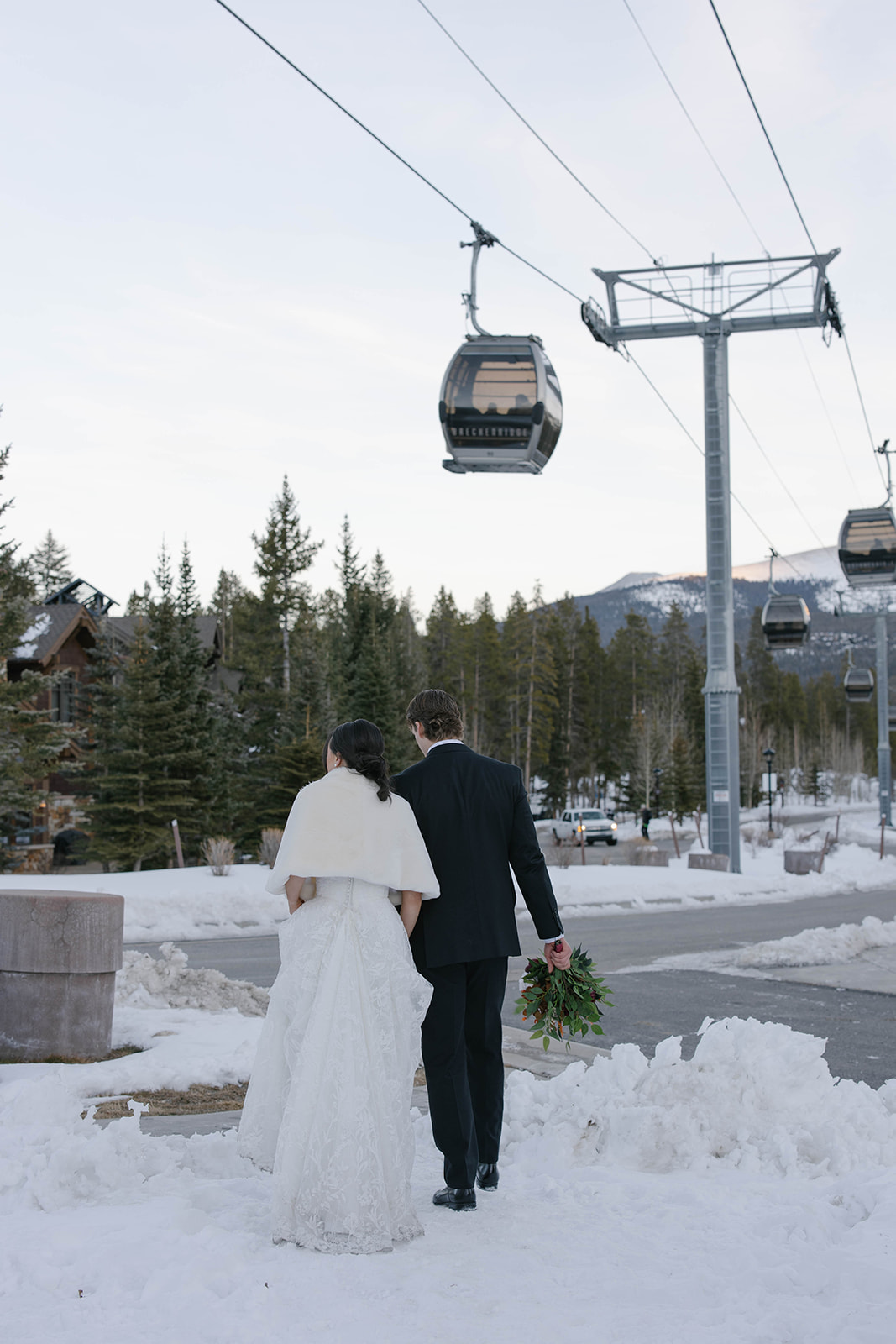 Bride and groom walking hand-in-hand through a snowy mountain town sidewalk beneath ski gondolas, capturing a winter Breckenridge elopement adventure.