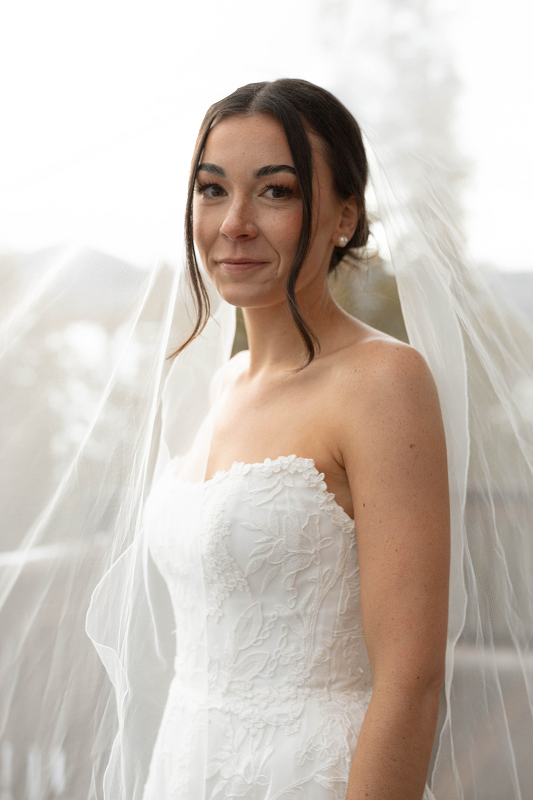 Bride smiling for the camera, sunlight glowing behind her and catching the edges of her veil, close-up portrait with shallow depth of field.