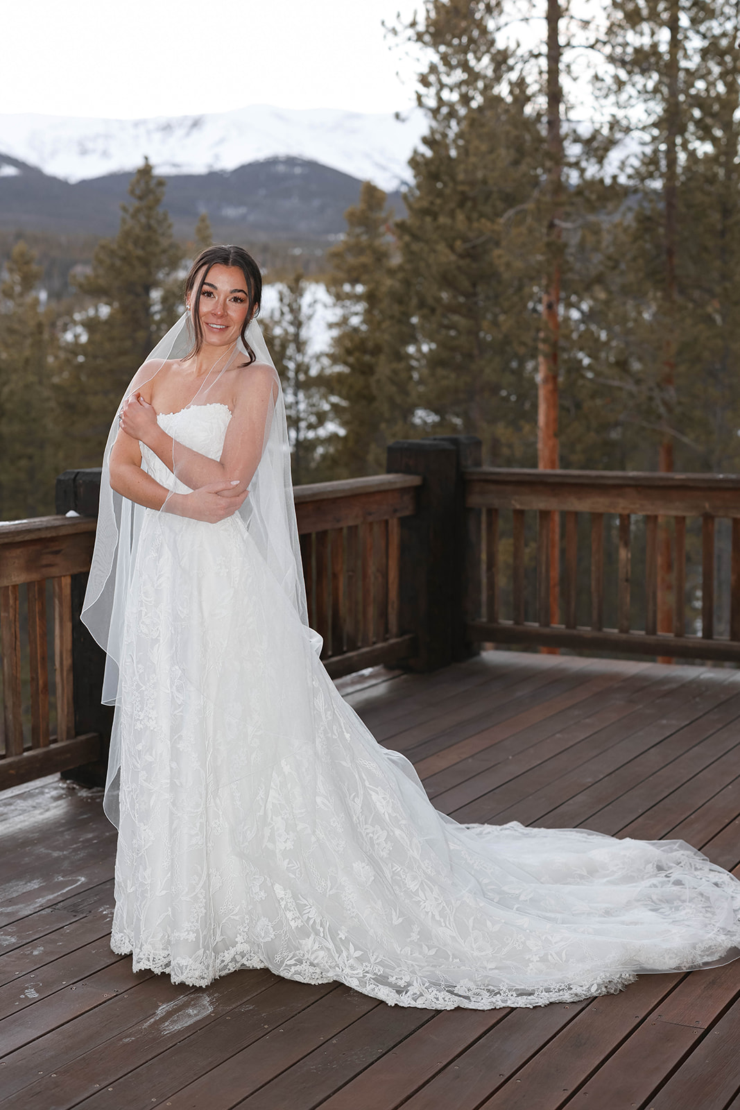Bride standing on a wooden deck in a lace wedding gown and veil, snow-dusted mountains and evergreen trees visible in the background.